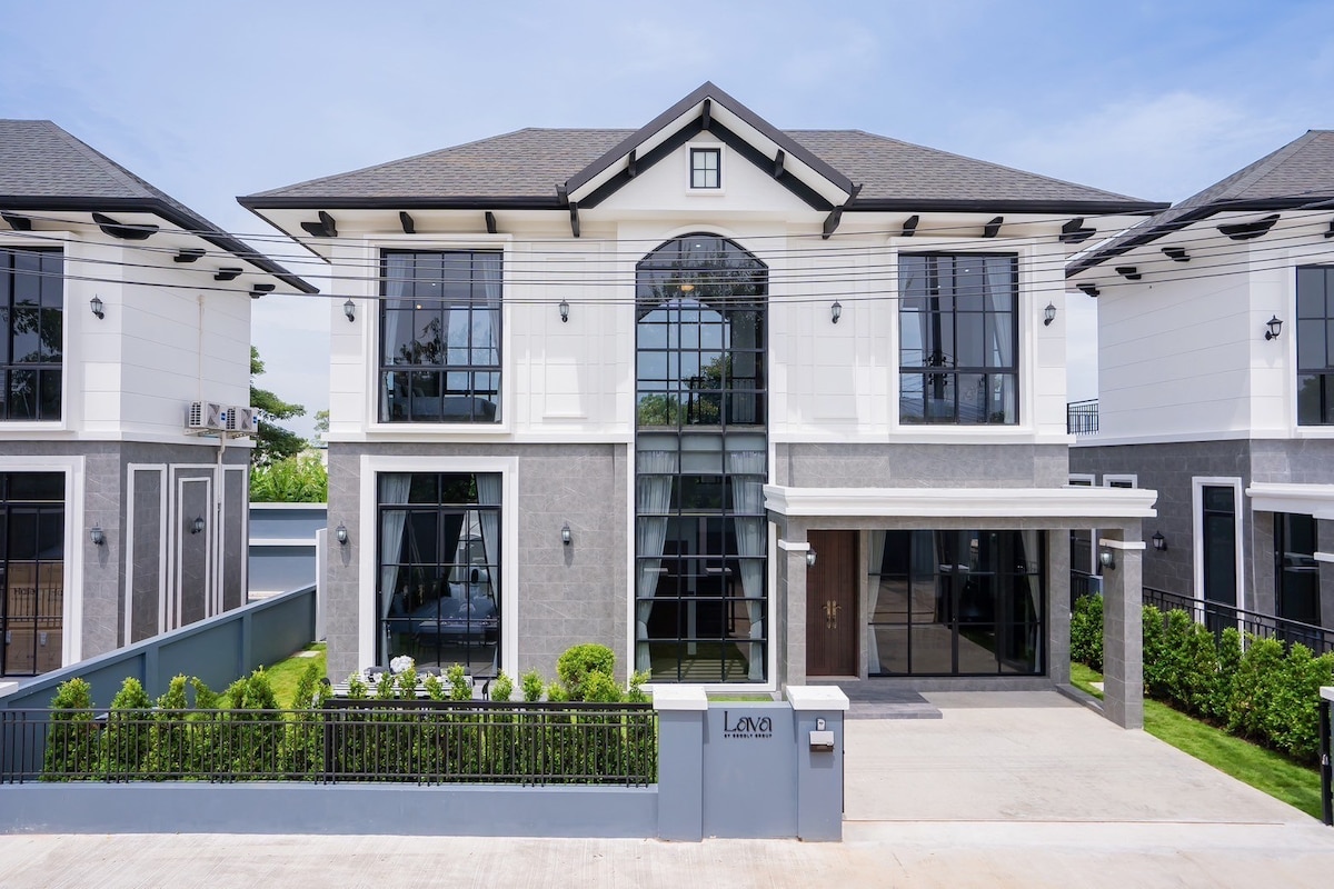 A modern two-story home featuring a symmetrical facade with large glass windows and a welcoming entrance. The exterior is characterized by a combination of gray stone and white paneling, complemented by manicured greenery along the front and side pathways.