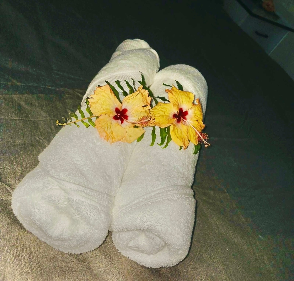 Two rolled white towels are arranged neatly on a textured surface, accented by yellow hibiscus flowers and green foliage placed on top. The setting suggests attention to detail and comfort.