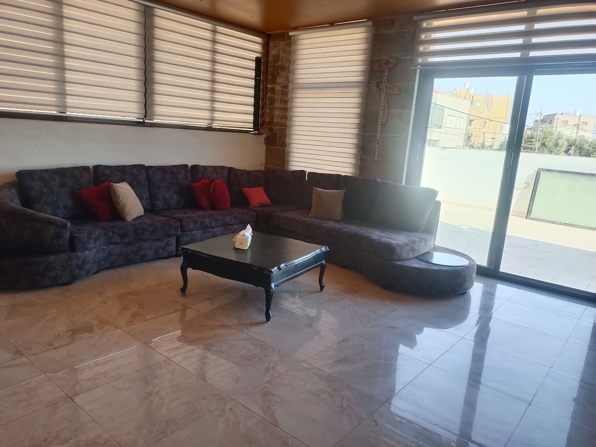 A spacious living area features a large, dark sectional sofa with decorative cushions. A sleek coffee table sits in front of the sofa, with large windows allowing natural light to brighten the room. Vertical blinds filter light, creating a calm ambiance.
