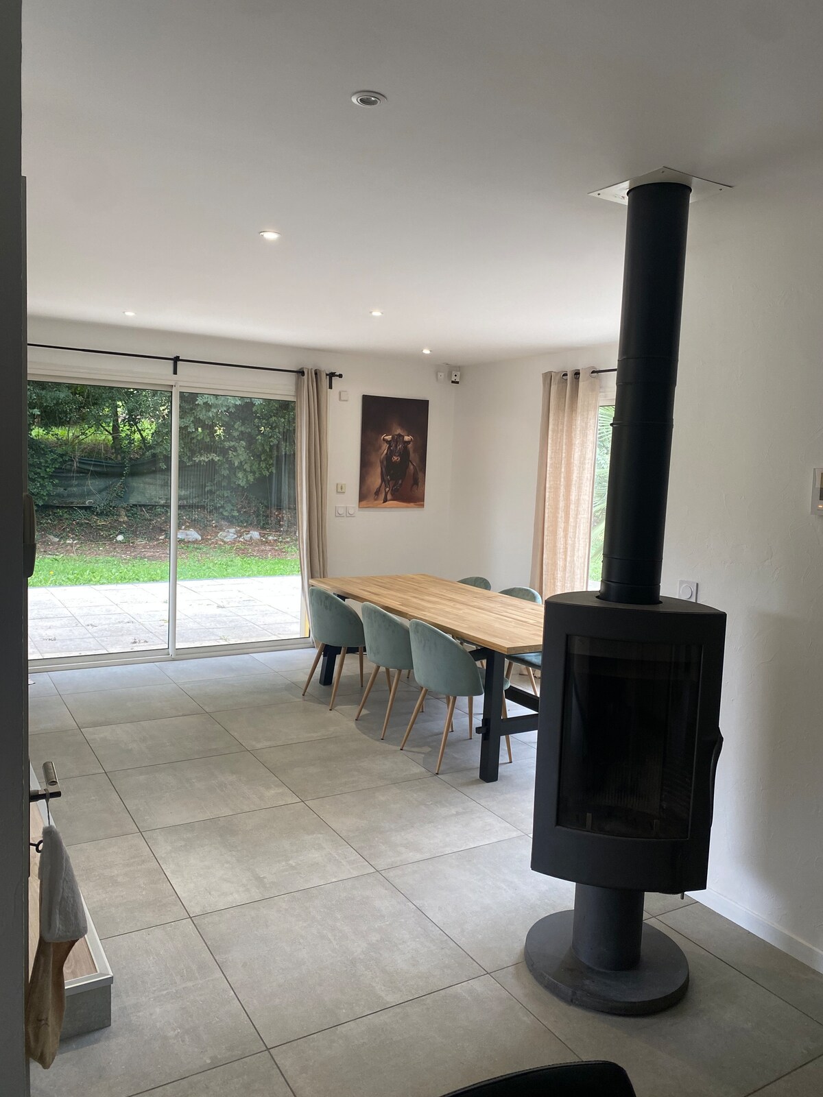 A bright dining area features a large wooden table surrounded by soft green chairs. A modern stove stands in the corner, providing warmth. Large glass doors lead to an outdoor space, allowing natural light to fill the room. Neutral tones accentuate the minimalist design.