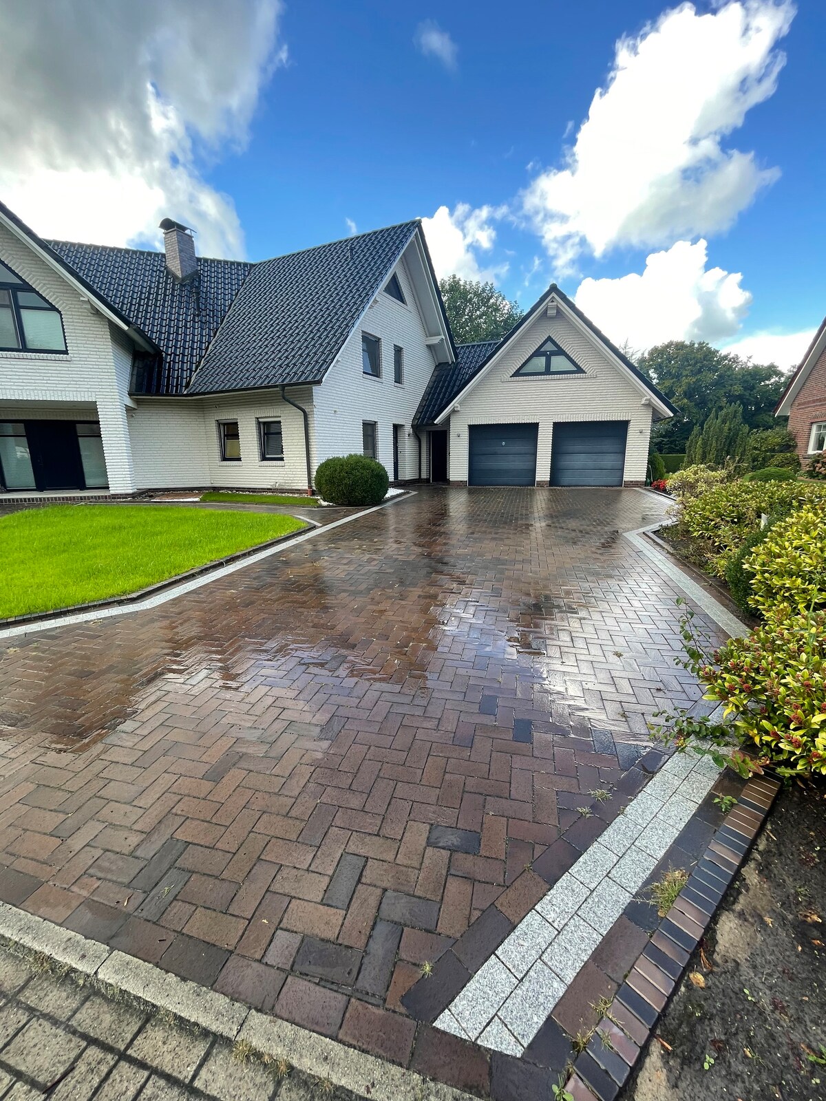 A modern residential exterior features a well-maintained driveway with a pattern of interlocking bricks, leading to a double garage. Lush green grass borders the pathway, and shrubs add natural elements, while the cloudy sky enhances the overall scene.