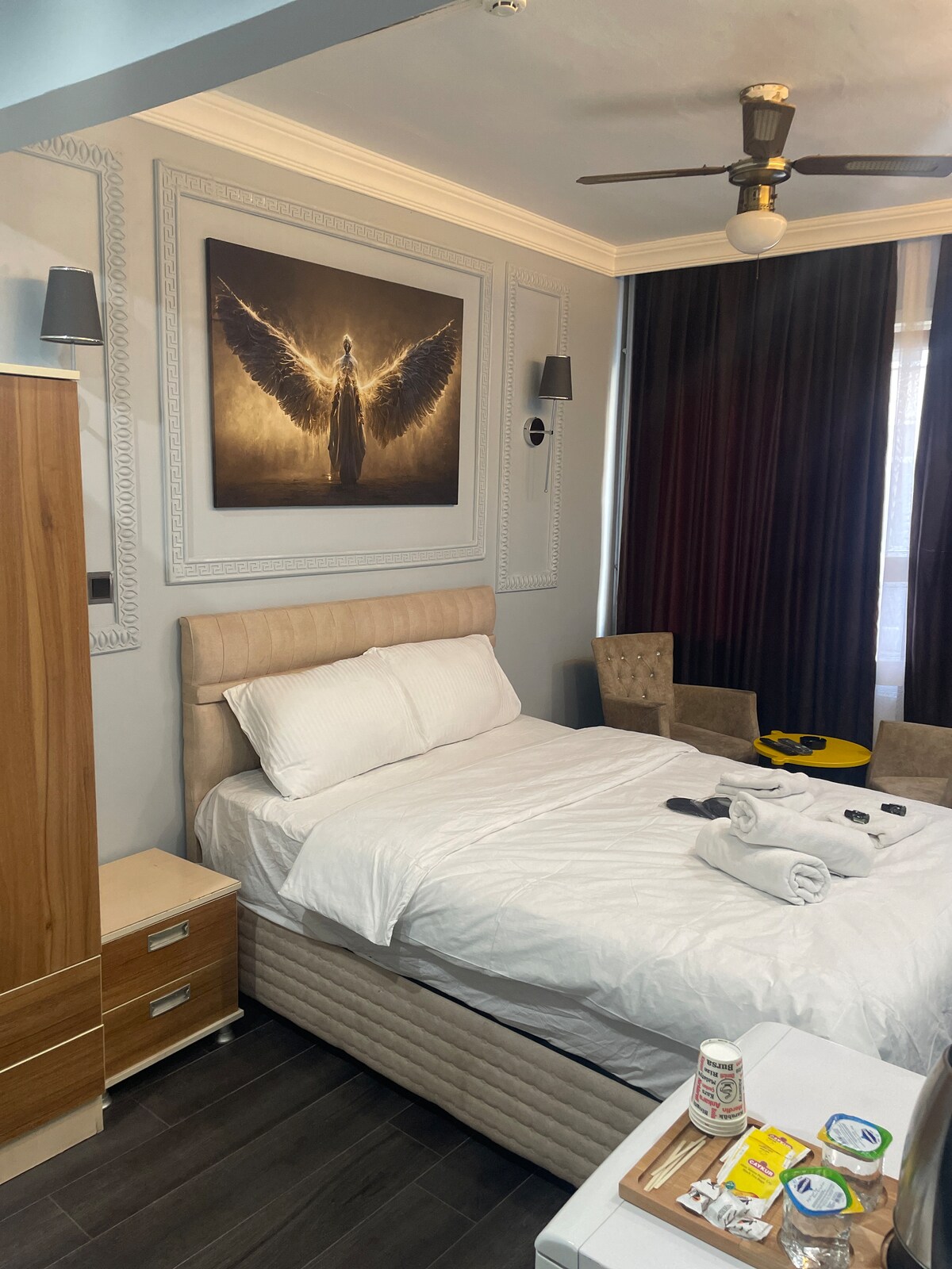 A spacious bedroom features a neatly made bed with white bedding and plush pillows. Two stylish armchairs sit beside a small round table. A large wall art piece depicting a figure with wings is prominently displayed above the bed, complemented by modern wall sconces.