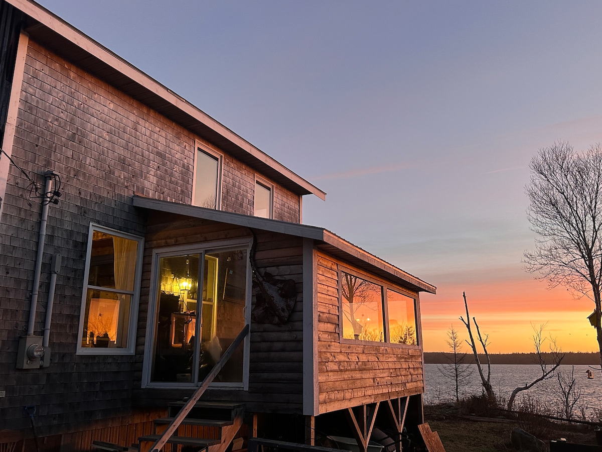 The exterior of a wooden house is captured during sunset, revealing large windows that reflect warm light. The water and sky are painted in soft hues of orange and pink. A deck leads to the water's edge, framed by trees in the distance.