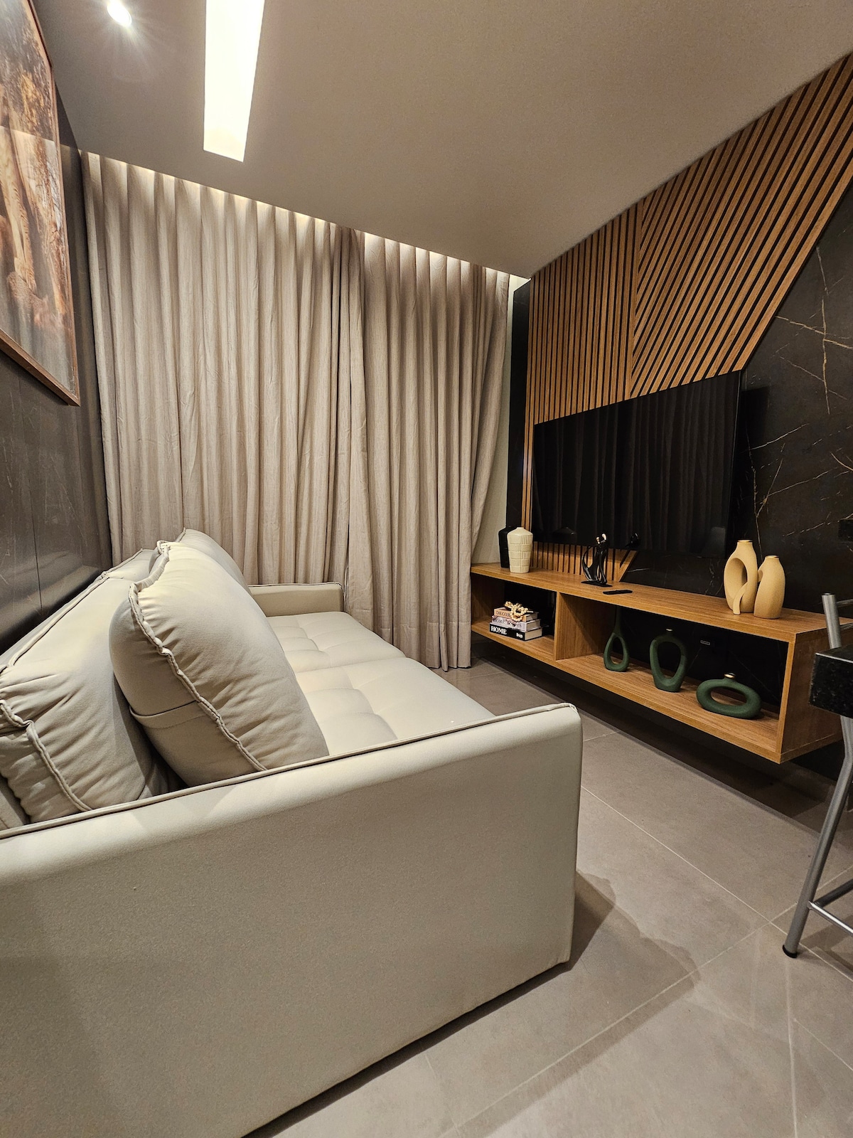A stylish living area showcases a plush light-colored sofa positioned against a textured wall. A mounted TV is complemented by minimalist shelving featuring decorative items. Floor-to-ceiling curtains frame the space, enhancing the cozy ambiance.