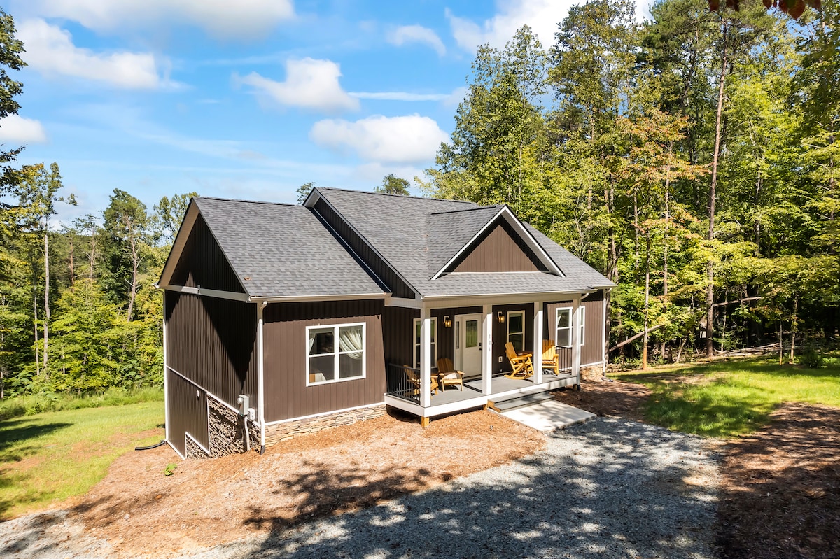 A modern single-story house is set among lush trees, featuring a covered porch with two rocking chairs. The exterior showcases dark siding and a well-maintained front yard. Natural light illuminates the space, creating a serene, inviting atmosphere.