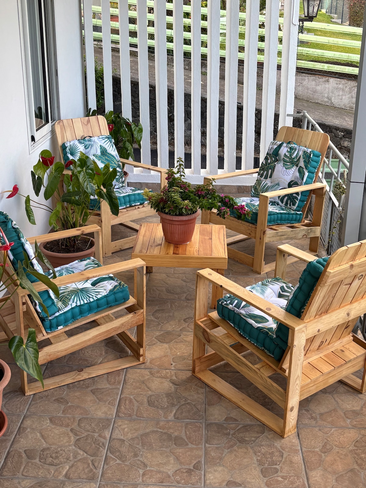 A cozy outdoor seating area is depicted with four wooden chairs, each featuring green patterned cushions. A small wooden table is positioned at the center, accompanied by potted plants that add a touch of greenery and life to the space.