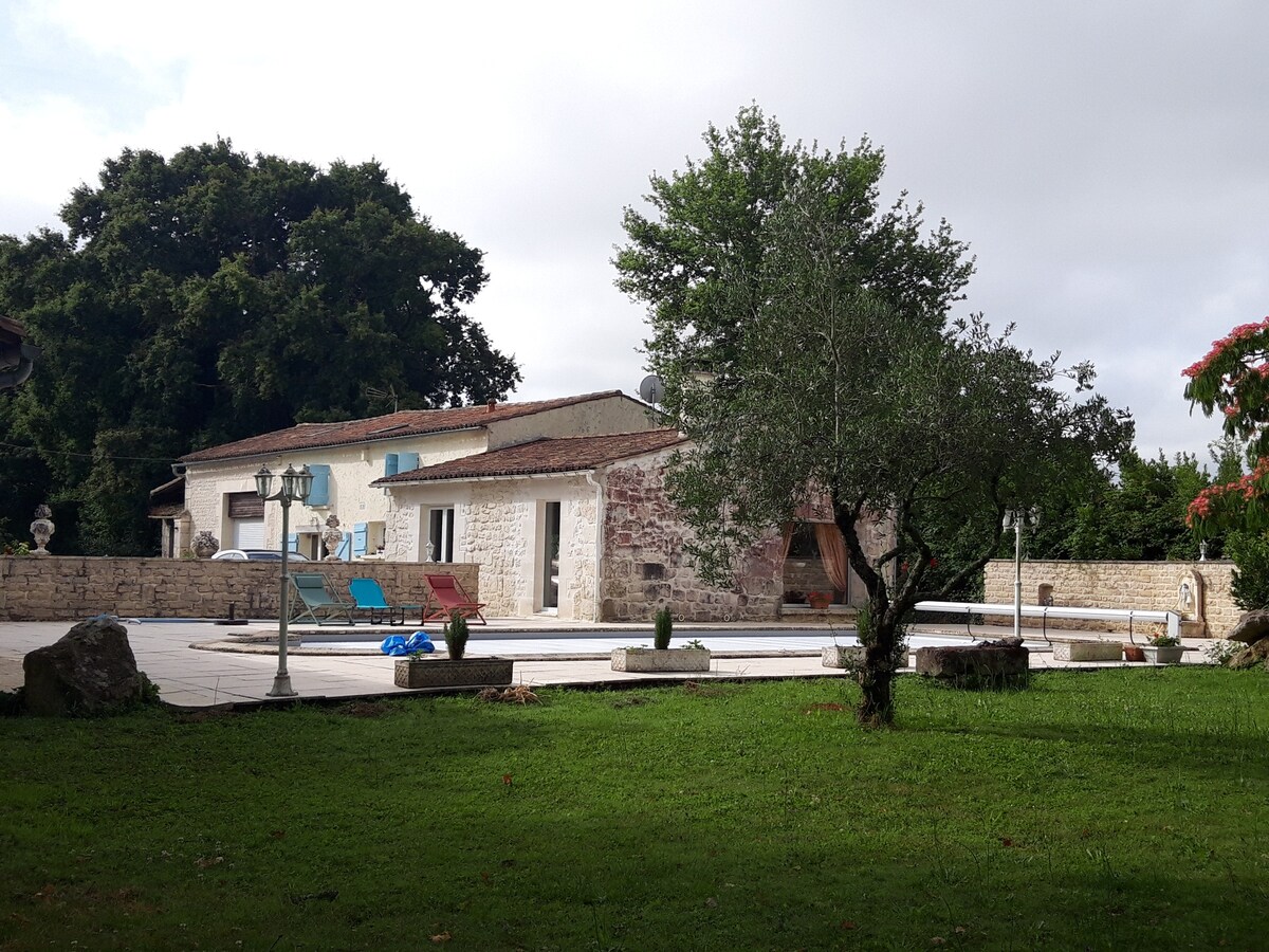 A charming stone building with blue shutters is surrounded by a lush green lawn. The spacious patio features two colorful lounge chairs beside a pool, complemented by decorative planters and an olive tree. A serene atmosphere is created by the nearby trees and nature.
