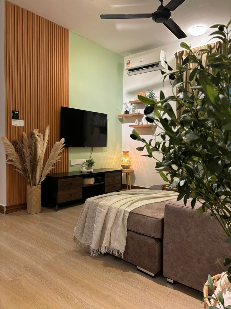 A serene living area features a cozy seating arrangement with a brown ottoman and a neatly draped blanket. A wall-mounted television is positioned above a dark wood console. Decorative plants and soft lighting add a calming touch, complemented by natural wood accents on the walls.