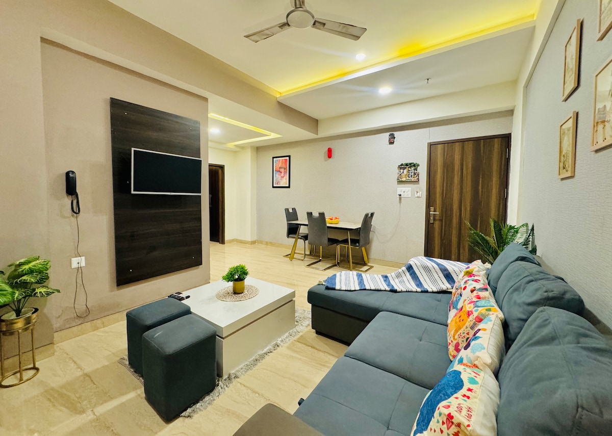 A spacious living area is presented, featuring a large sectional sofa adorned with decorative pillows. A coffee table occupies the center, while a mounted television adds a modern touch. Dining space is visible in the background, accompanied by light wood furniture and warm ambient lighting.