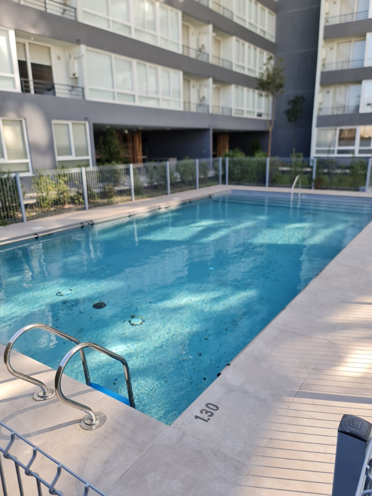 An outdoor swimming pool is situated within a fenced area, featuring clear blue water and a shallow entry. Surrounding greenery adds a natural element, and the pool is bordered by a smooth, tiled deck. Sunlight creates reflections on the water's surface.