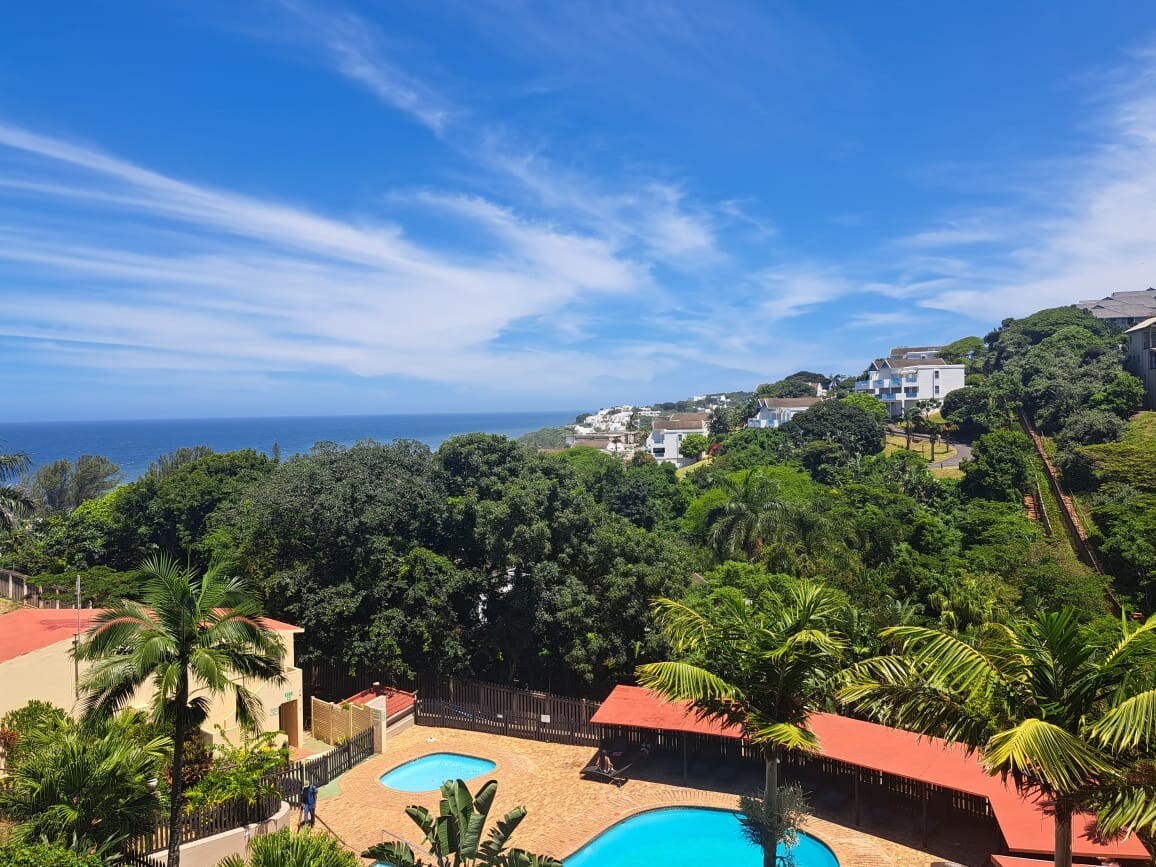 A scenic view captures a bright blue sky stretching over a tranquil coastal landscape. Two inviting swimming pools are surrounded by lush greenery. The soft contours of the hills in the background complement the serene atmosphere of the area.