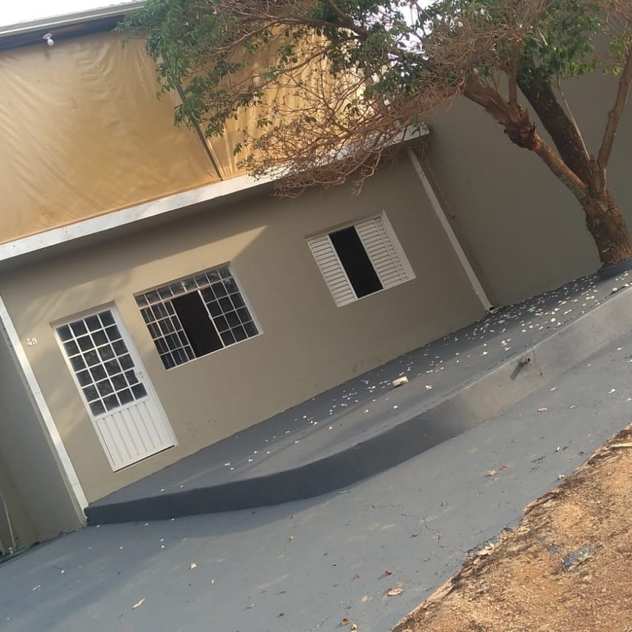 The exterior of a single-story house is showcased, featuring a muted color scheme. A large tree shades the area, and a small concrete ramp leads to the entrance. Windows are partially covered with shutters, providing a glimpse of the interior.