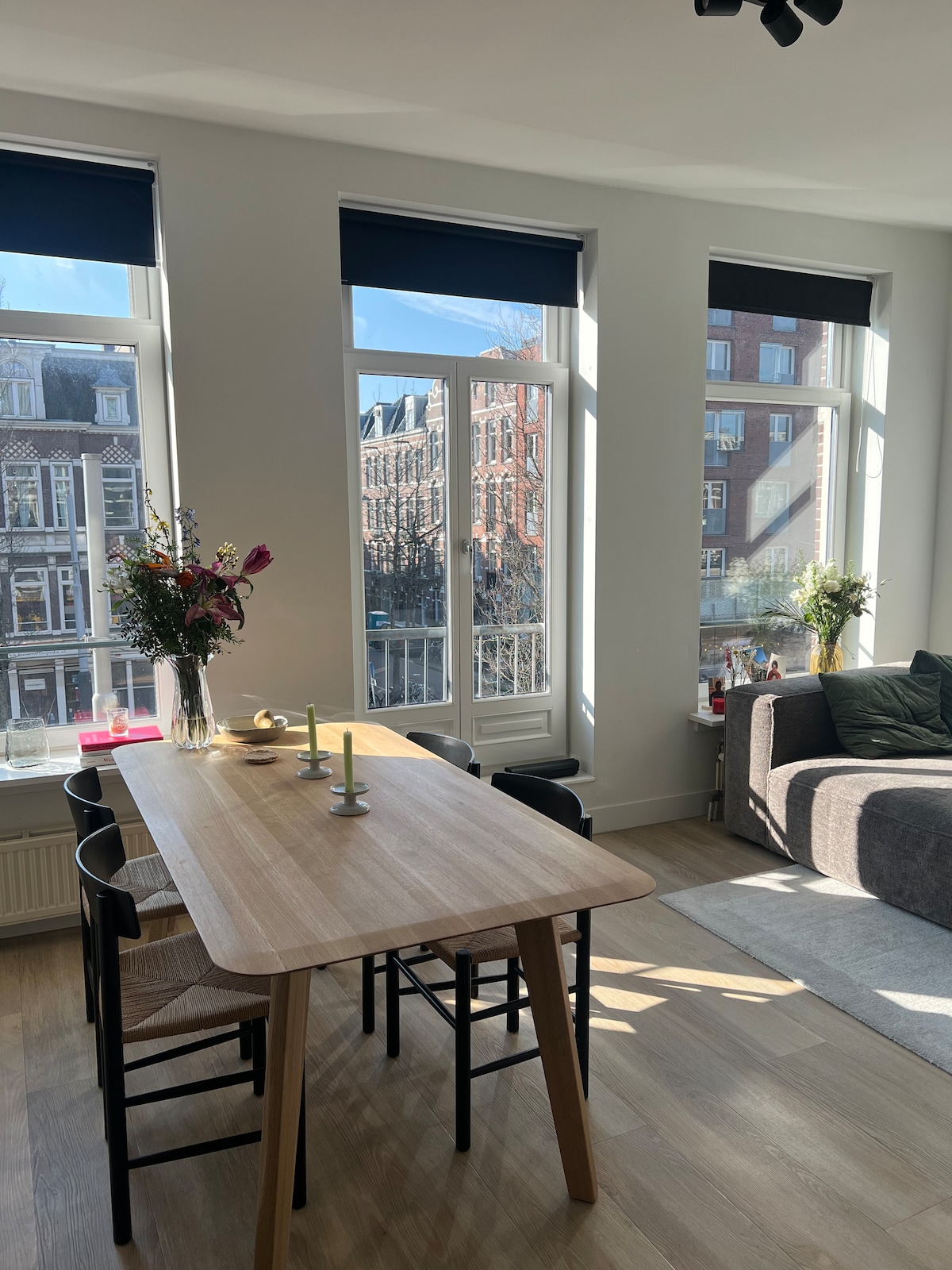 A dining area features a light wooden table surrounded by black chairs, positioned near large windows that allow natural light to fill the space. A vase with flowers and a few decorative items sit on the table, while a cozy sofa is visible in the background.