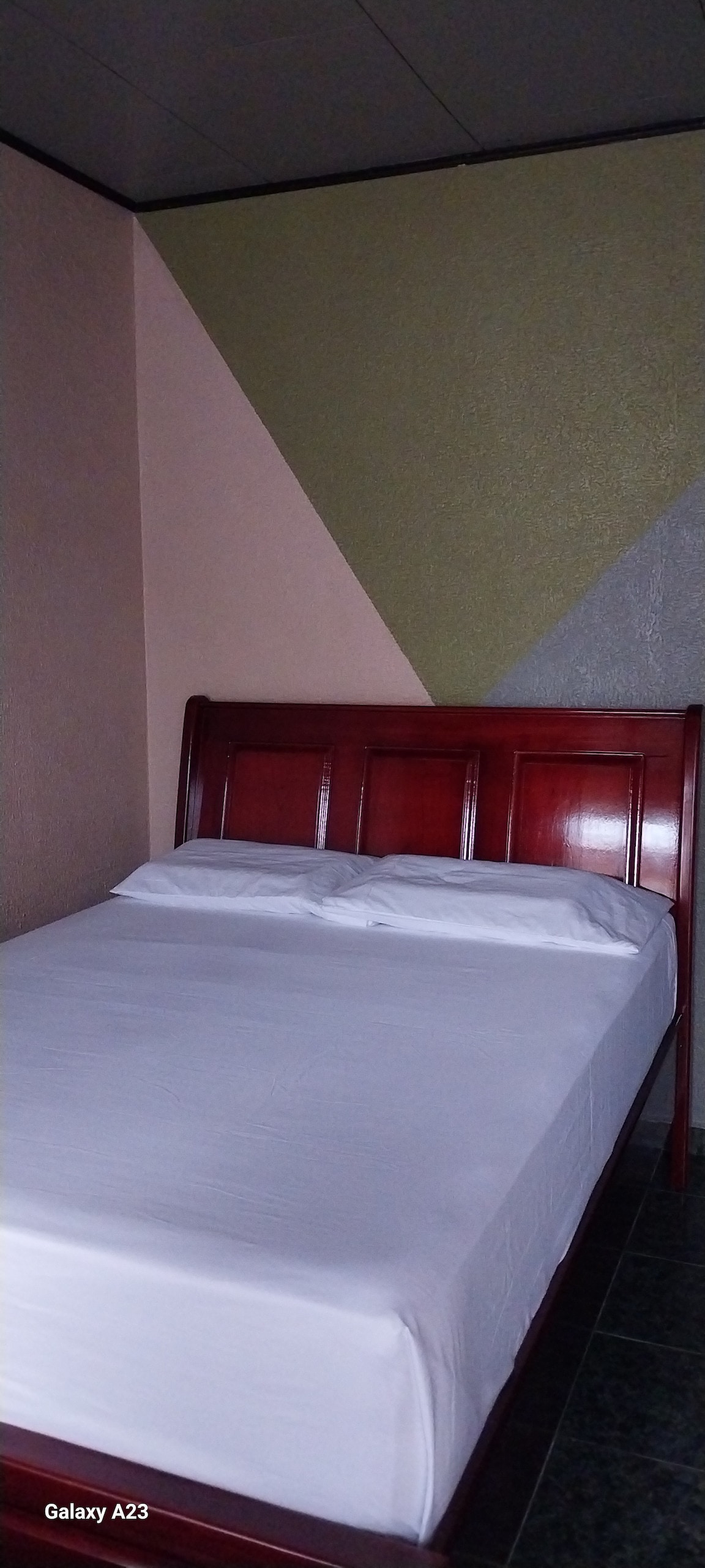 The image shows a wooden bed with a neatly made white bedspread. The wall behind features a geometric design in muted colors, creating a simple yet functional sleeping area. Soft lighting enhances the tranquility of the room.