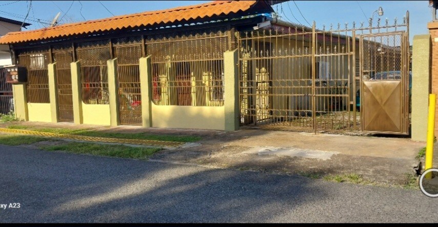 The exterior view features a gated entrance with a gold-toned fence and a large gate. A well-maintained pathway leads to the entrance of the building, surrounded by a green lawn. The roof showcases a traditional design, complementing the overall structure.
