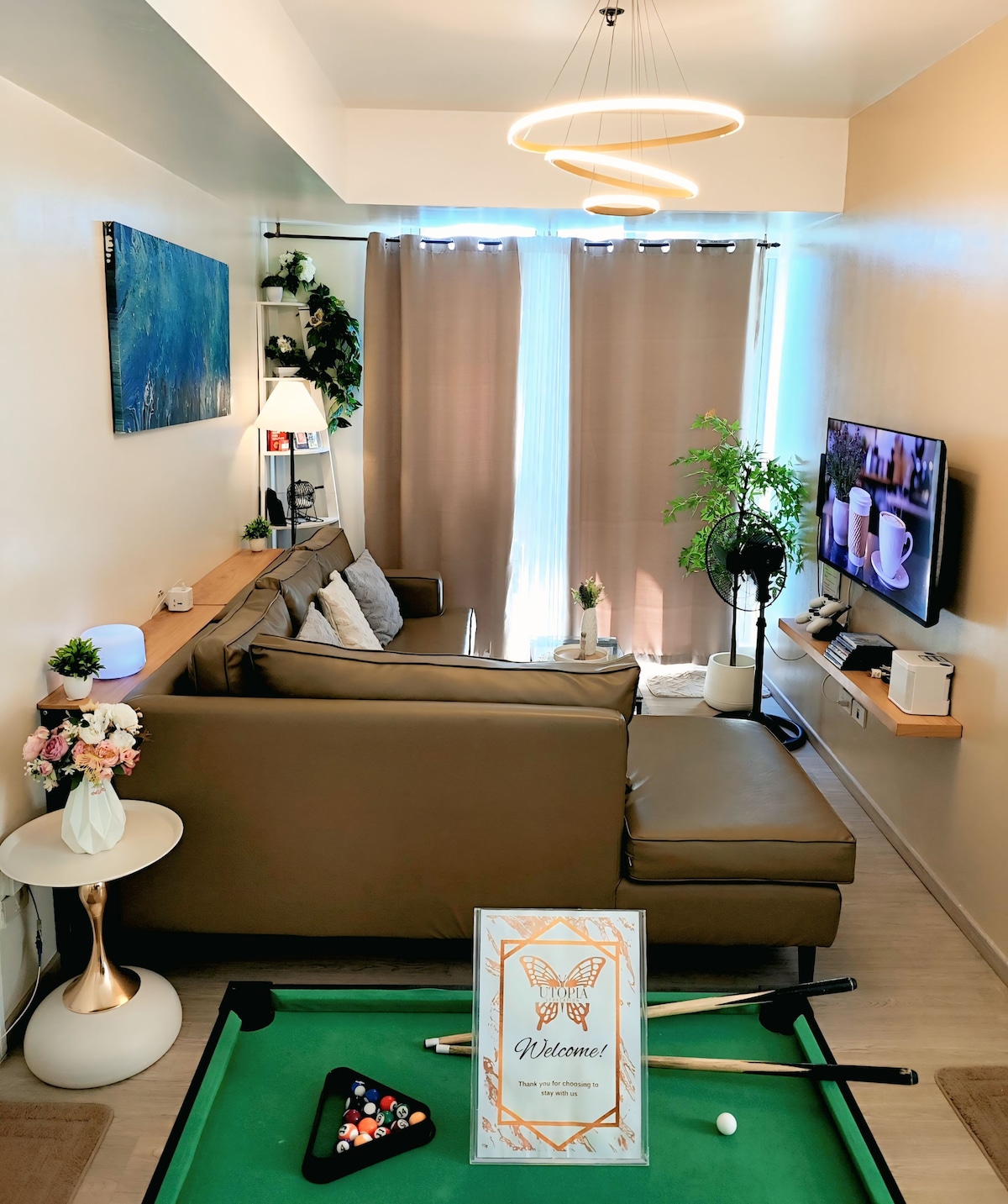 The living area displays a comfortable brown sectional sofa with decorative cushions, positioned next to a wall-mounted television. A mini billiards table is seen in the foreground, with a welcome sign and colorful pool balls. Natural light enters through large windows, softened by sheer curtains.