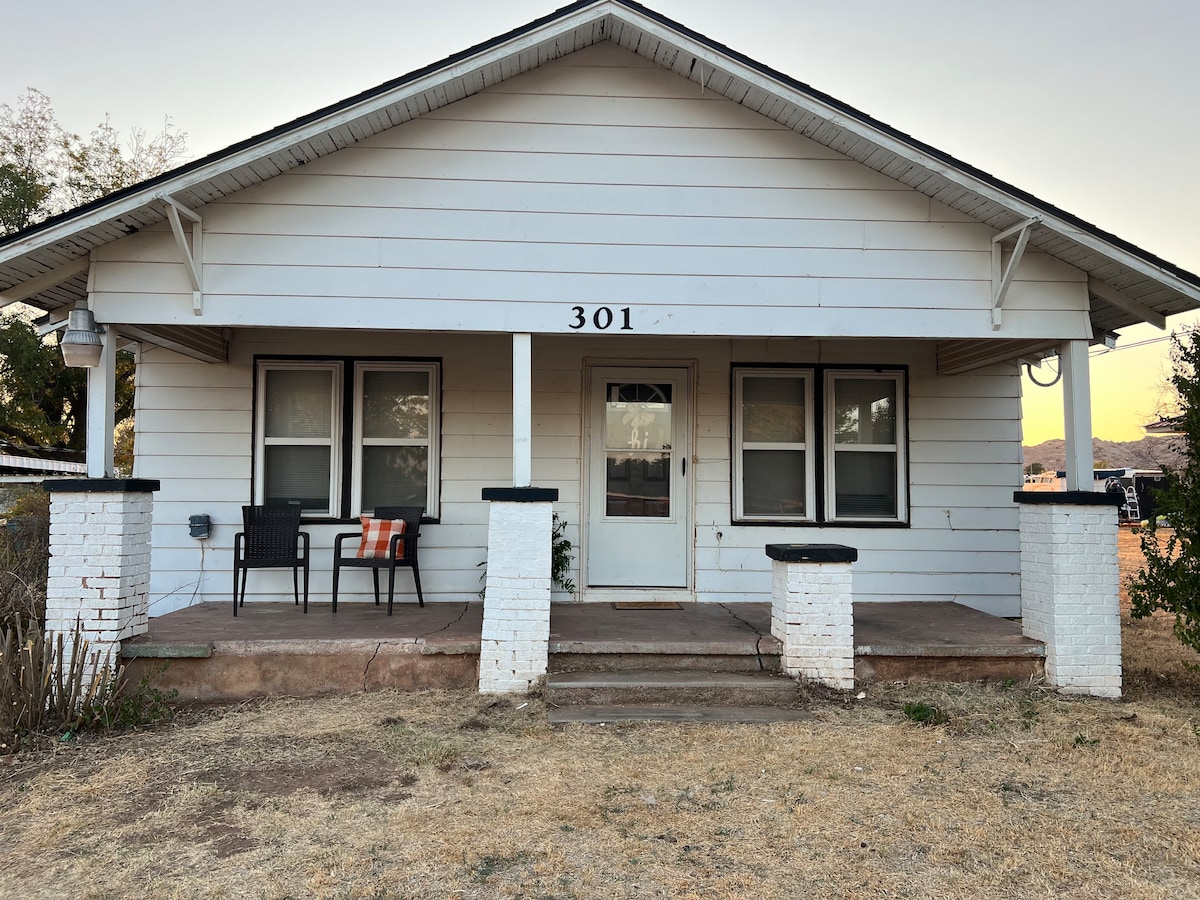 A covered front porch is showcased, featuring two chairs that invite relaxation. Decorative white brick columns frame the entrance, while large windows provide ample natural light. The surrounding area is a blend of brown grass and a clear dusk sky.