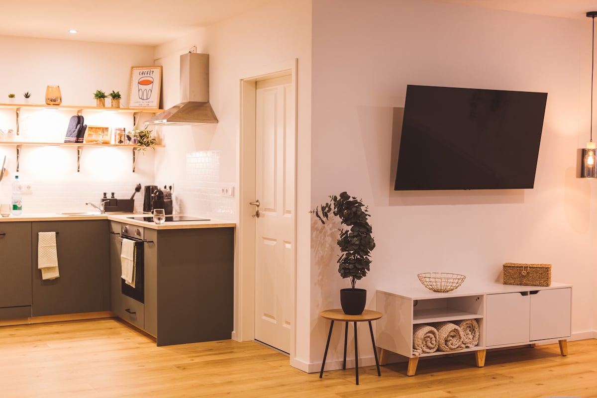 A modern kitchen area features sleek cabinetry and a spacious countertop with kitchen appliances. A flat-screen television is mounted on the wall opposite a small side table adorned with a potted plant. Natural light brightens the wooden flooring, enhancing the welcoming atmosphere.
