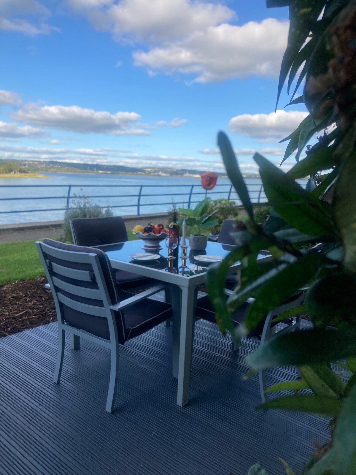 A dining area is positioned on a patio overlooking the waterfront, featuring a sleek table set with dinnerware and floral decor. Comfortable chairs surround the table, with greenery framing the scene, capturing the tranquil outdoor setting against a backdrop of open skies.