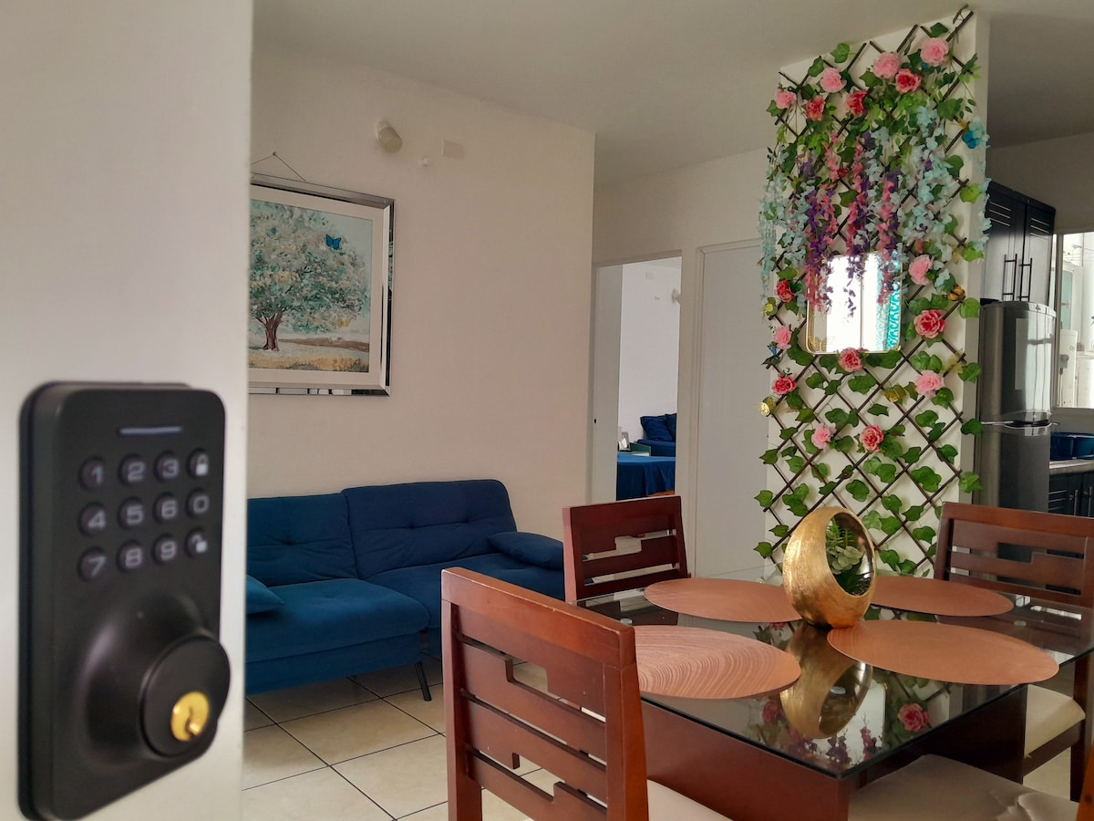 A modern living area showcases a blue sofa and a dining table with a circular centerpiece. A decorative wall adorned with greenery and flowers adds a fresh accent, while a keypad lock is visible near the entrance.