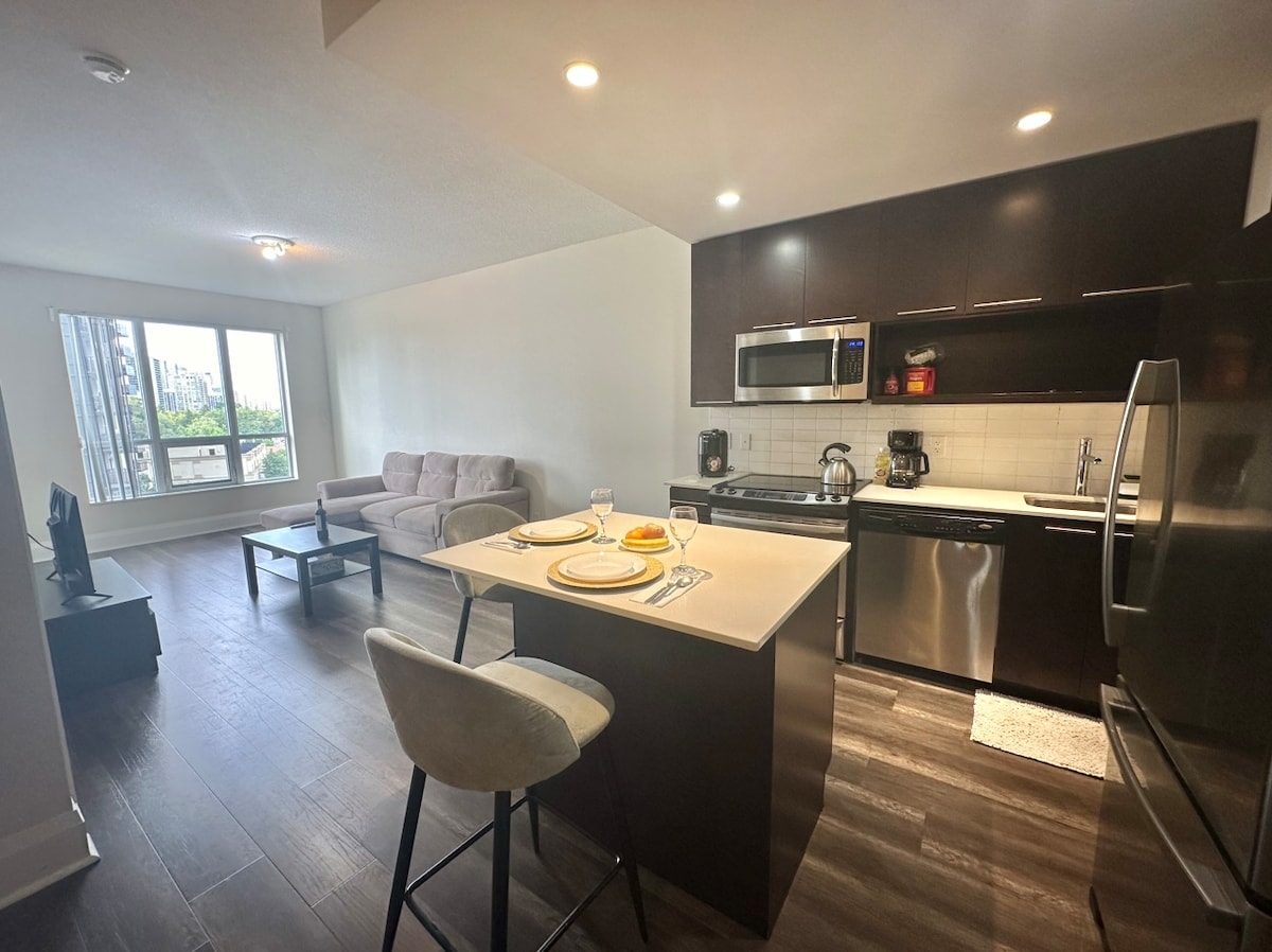 A modern kitchen is featured with stainless steel appliances and a breakfast bar. A comfortable living area is visible, furnished with a soft sofa and a coffee table, complemented by large windows that allow natural light to fill the space.