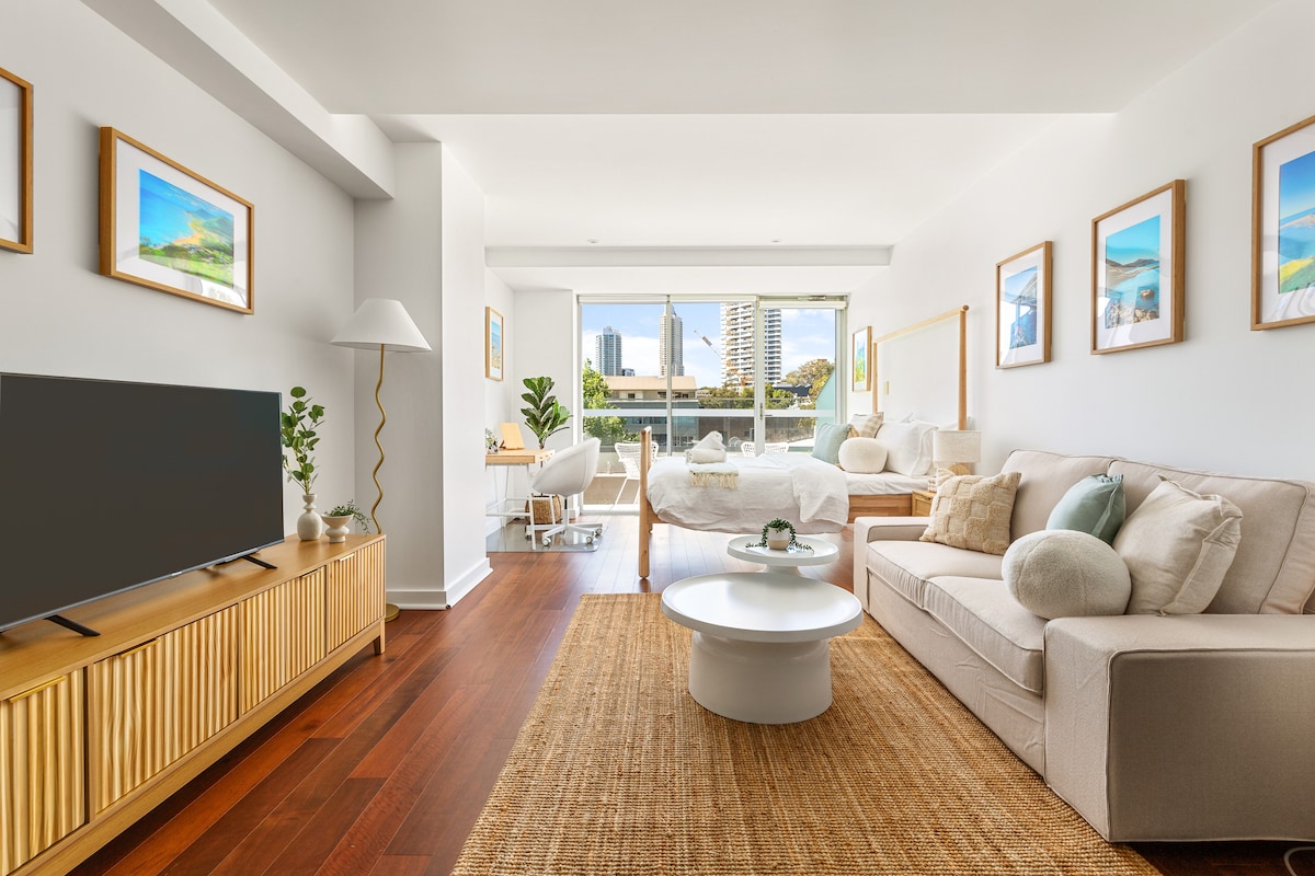 The open layout of the studio includes a cozy living area with a light-colored sofa and a low coffee table on a natural fiber rug. A dining space is visible beside large windows, allowing natural light to fill the room. Framed artworks adorn the walls.