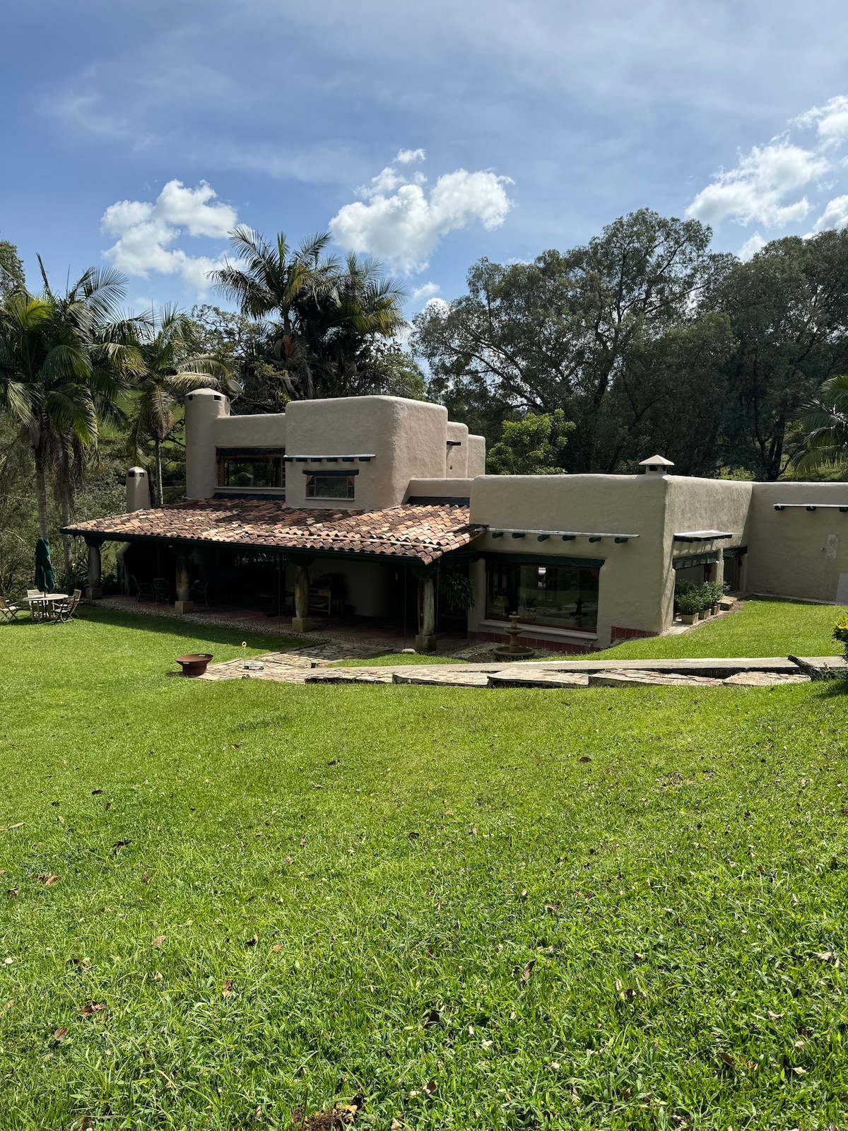 The exterior of the charming house is showcased, featuring a unique architectural design with a terracotta roof and textured walls. Lush greenery surrounds the property, with trees and a well-maintained lawn visible. Outdoor seating and a pathway enhance the inviting atmosphere.