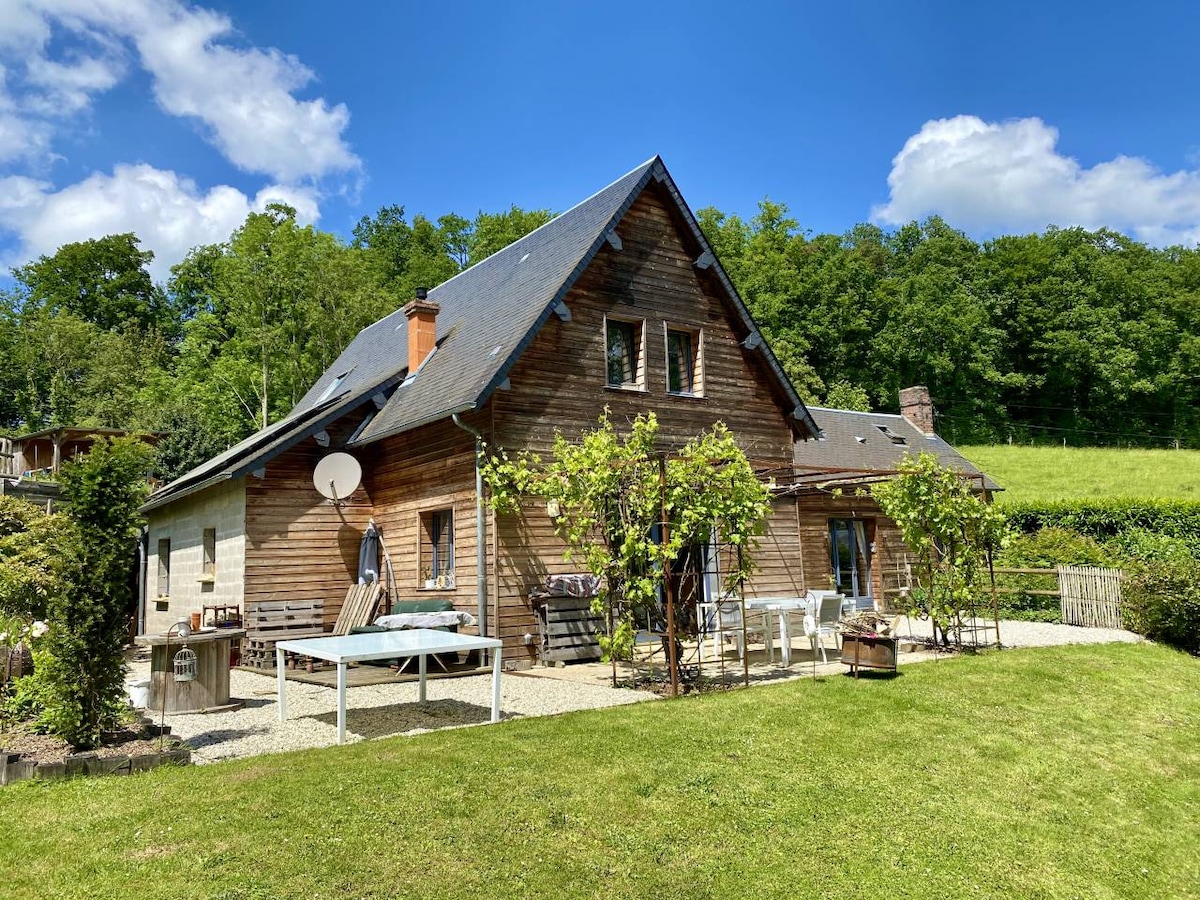A wooden house is nestled against a backdrop of green foliage. The exterior features multiple outdoor seating areas, including a spacious table, and is surrounded by a well-kept lawn. Sunlight brightens the scene, and blue skies add a serene quality to the setting.