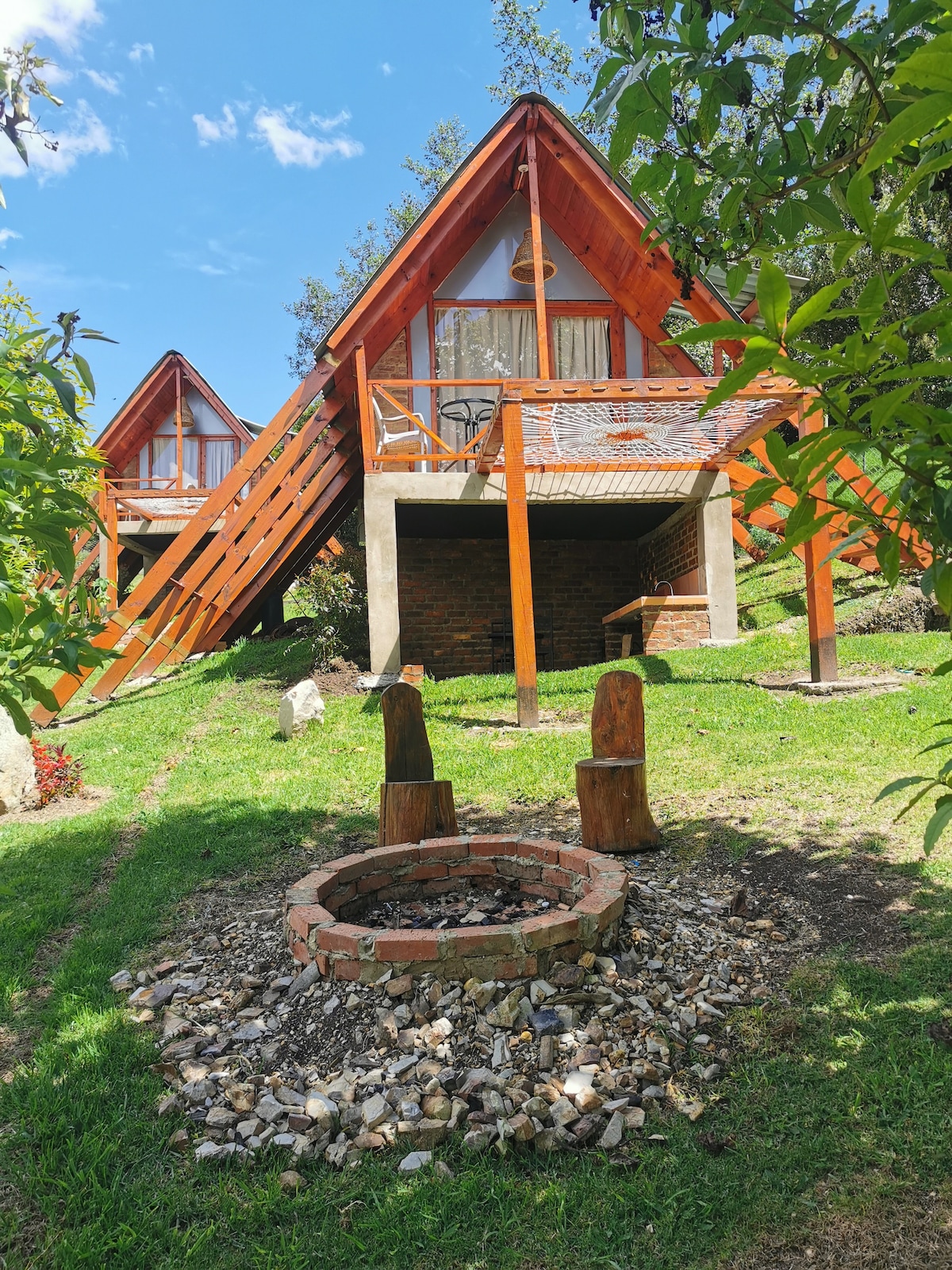 The exterior of a wooden cabin is surrounded by lush greenery, featuring a stone fire pit encircled by seating. The structure displays a gabled roof with large glass windows, while a wooden deck provides access to an elevated entryway. Natural light floods the area.