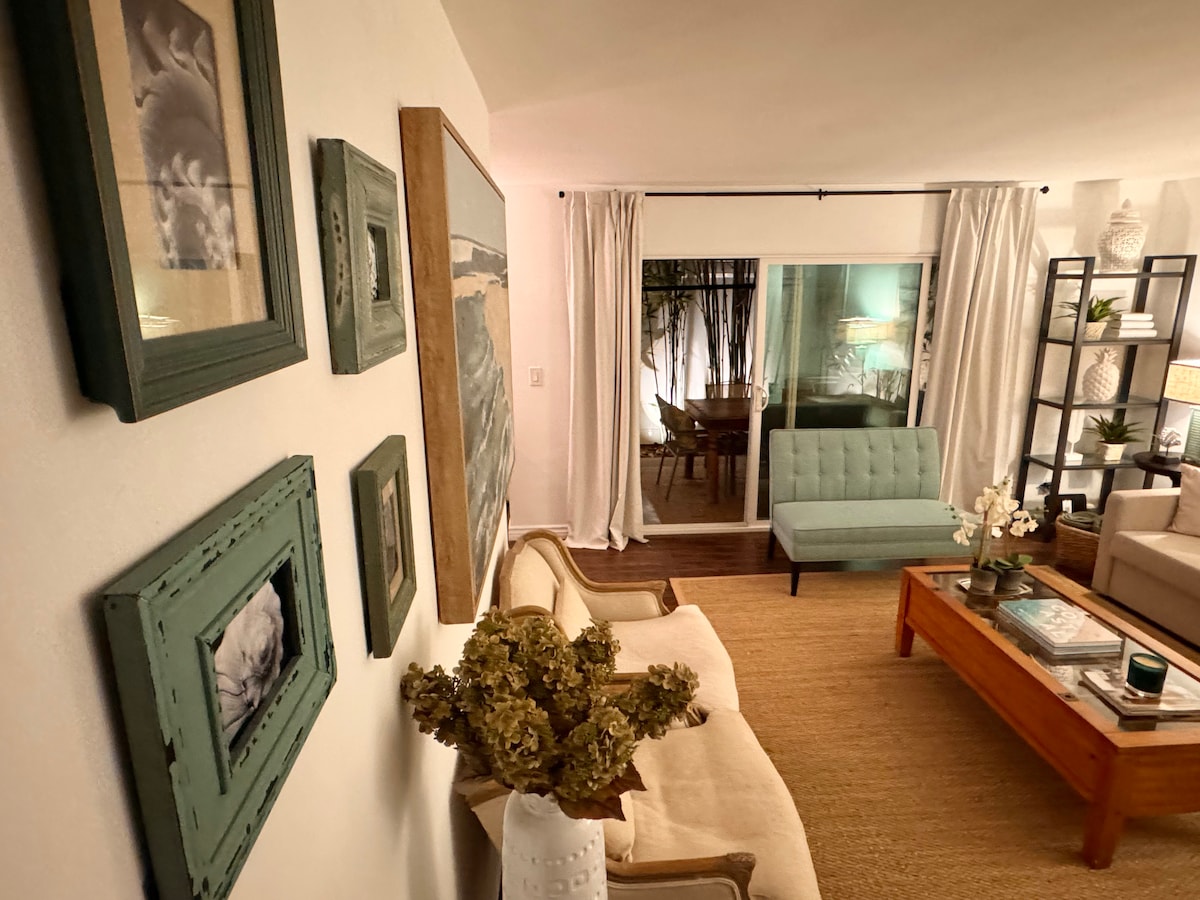 A comfortable living room is featured, showcasing a neutral sofa and a mint green chair. Artwork with natural themes adorns the walls, complemented by a decorative plant arrangement in a white vase. Natural light filters through sheer curtains, illuminating the space.