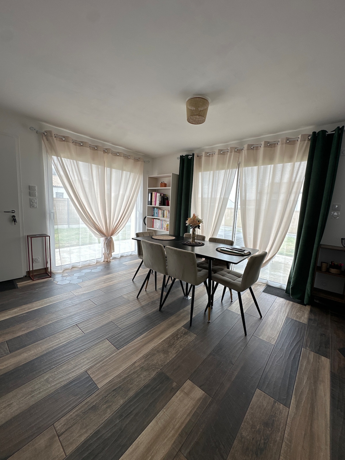 A bright and airy dining area is presented with a modern table and four chairs. Natural light filters through sheer curtains, enhancing the spacious feel. A bookcase filled with books is visible, along with a glimpse of greenery outside the window.