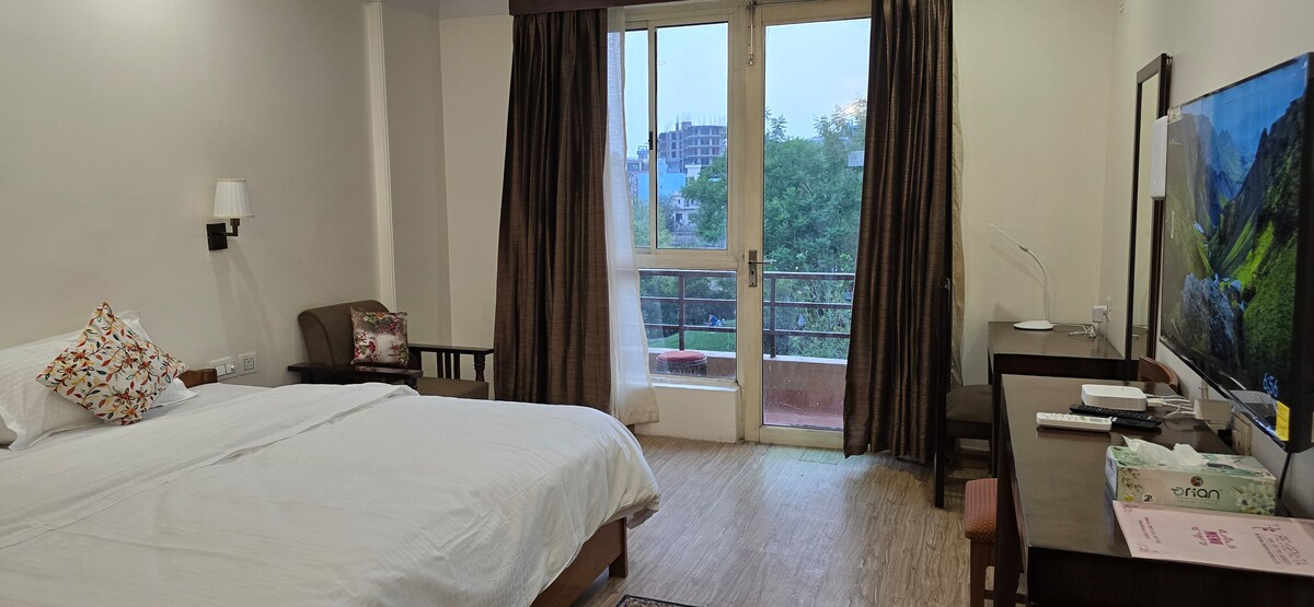 A comfortable bedroom is equipped with a spacious bed and a seating area featuring an armchair and a small table. A large window offers a view of the greenery outside, while a TV is positioned on a wooden console nearby.