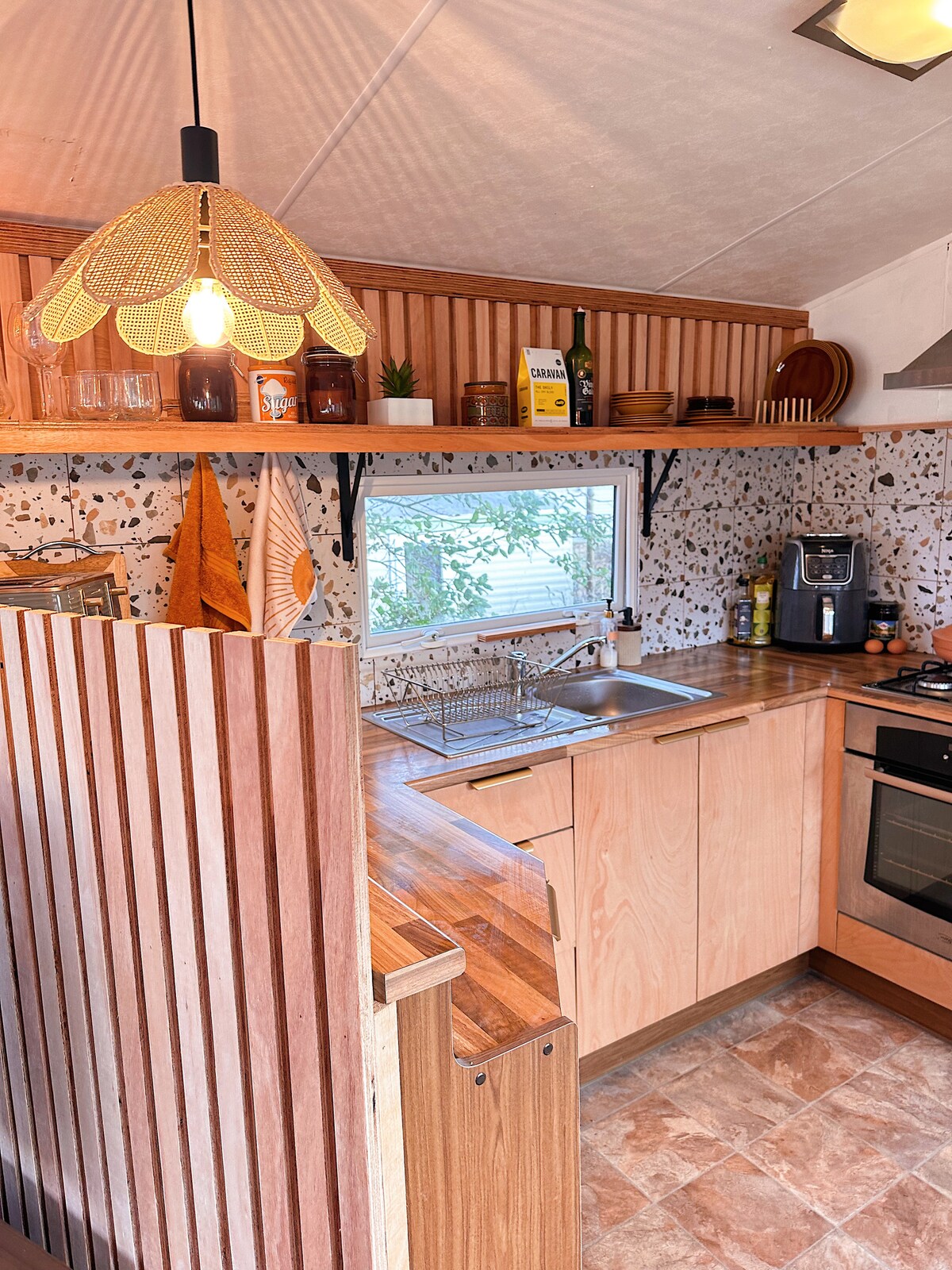 The kitchen area features natural wood cabinetry and a textured backsplash, accented by warm lighting from a decorative pendant lamp. A window allows natural light and offers views of the surrounding trees, while modern appliances are neatly arranged on the countertop.