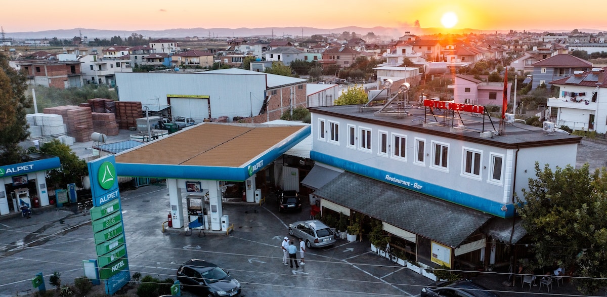 An aerial view captures a gas station and a restaurant area at sunset. The gas station features a prominent canopy and signage. Surrounding structures and vehicles are visible, with a backdrop of a sprawling town and distant mountains.