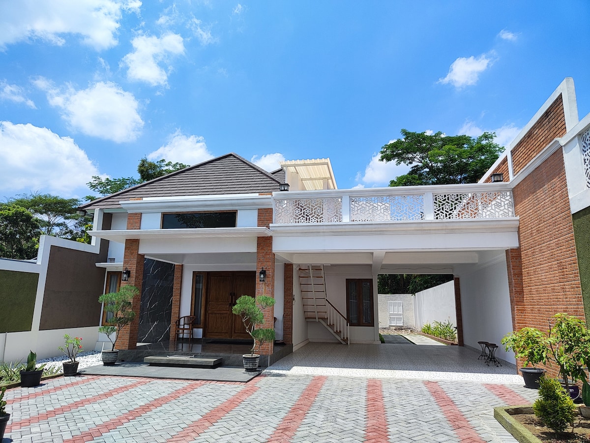 The exterior of a modern two-story homestay is showcased, featuring a combination of brick and white walls. A spacious entryway leads to a staircase, while potted plants enhance the outdoor area. The blue sky is visible above, providing a bright and open atmosphere.