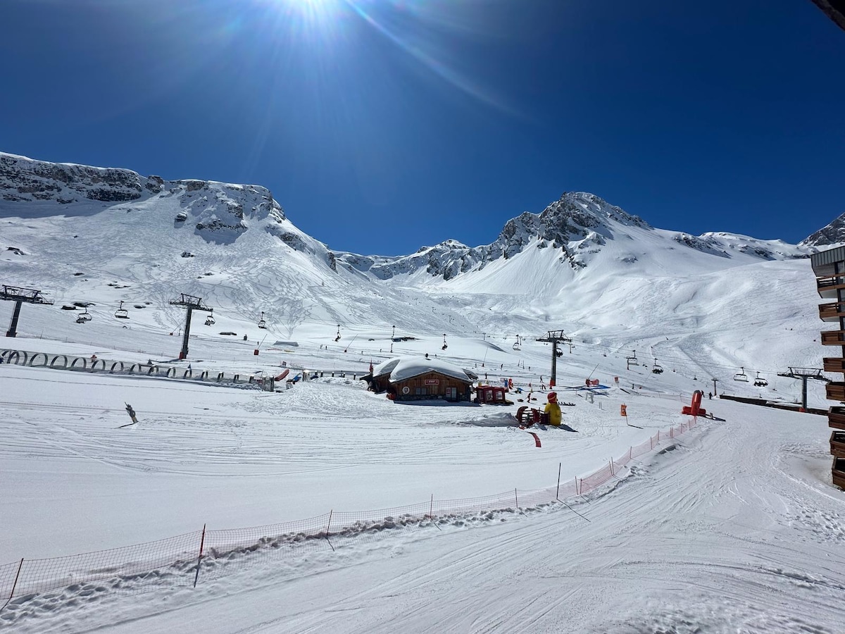 Snow-covered slopes extend towards distant mountains under a clear blue sky, with a sun casting bright light across the scene. Ski lifts and ski runs are visible, alongside a lodge situated on the terrain, providing access to the ski area.