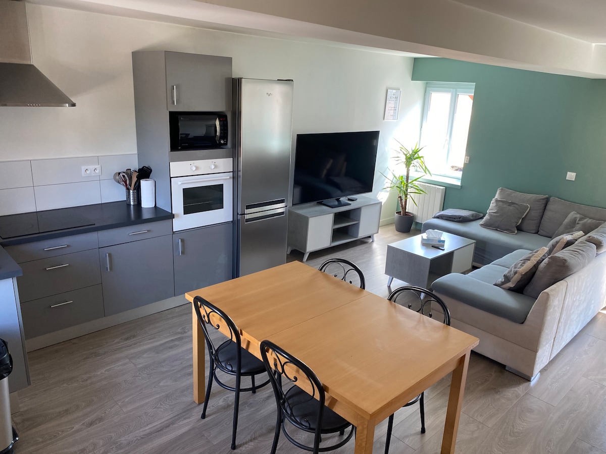 An open living area features a modern kitchen with gray cabinets and stainless steel appliances. A light-colored dining table with four black chairs is positioned beside a comfortable gray sofa. A wall-mounted television and a potted plant enhance the inviting atmosphere.