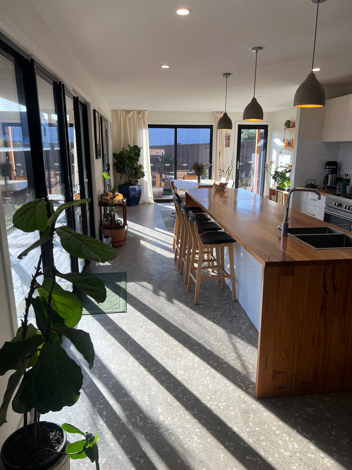 An open-plan kitchen and dining area features a long wooden island with seating for four. Tall glass doors allow natural light to flood the space, highlighting indoor plants and a cozy atmosphere. Pendant lights hang above, adding a touch of modern design.