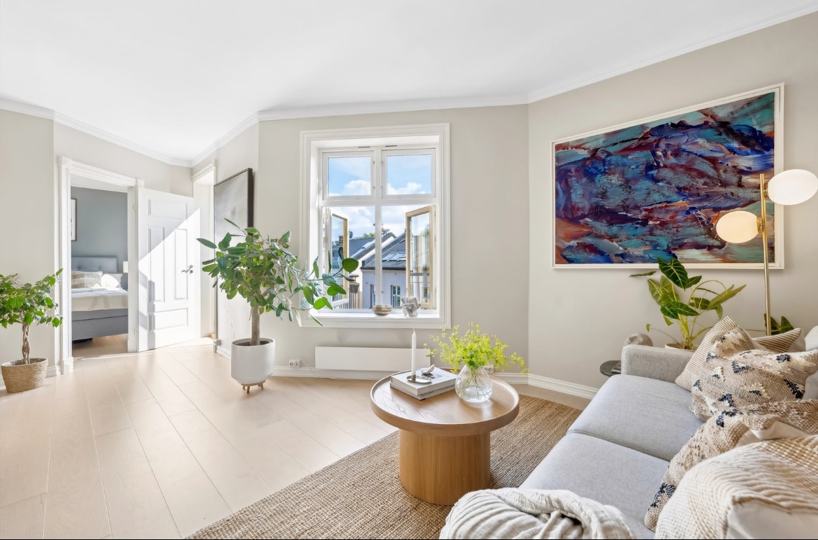 A light-filled living area showcases a gray sofa complemented by a round wooden coffee table. Large windows open to the outside, allowing ample natural light. Lush greenery and a vibrant wall art piece add visual interest to the space. Double doors lead to another room.
