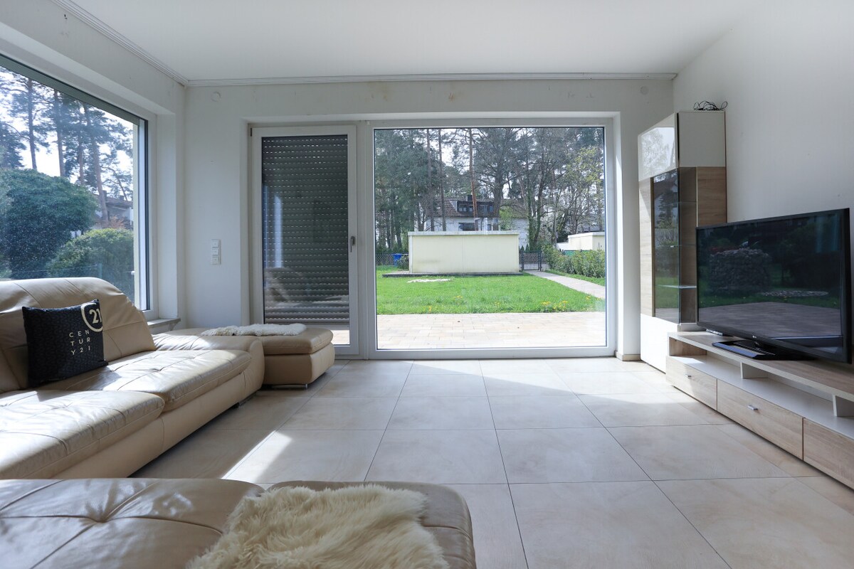 A well-lit living room features a spacious sectional sofa and large windows that provide views of the outdoor space. A television is positioned against one wall, while neutral-colored tiles cover the floor, enhancing the room's airy feel.