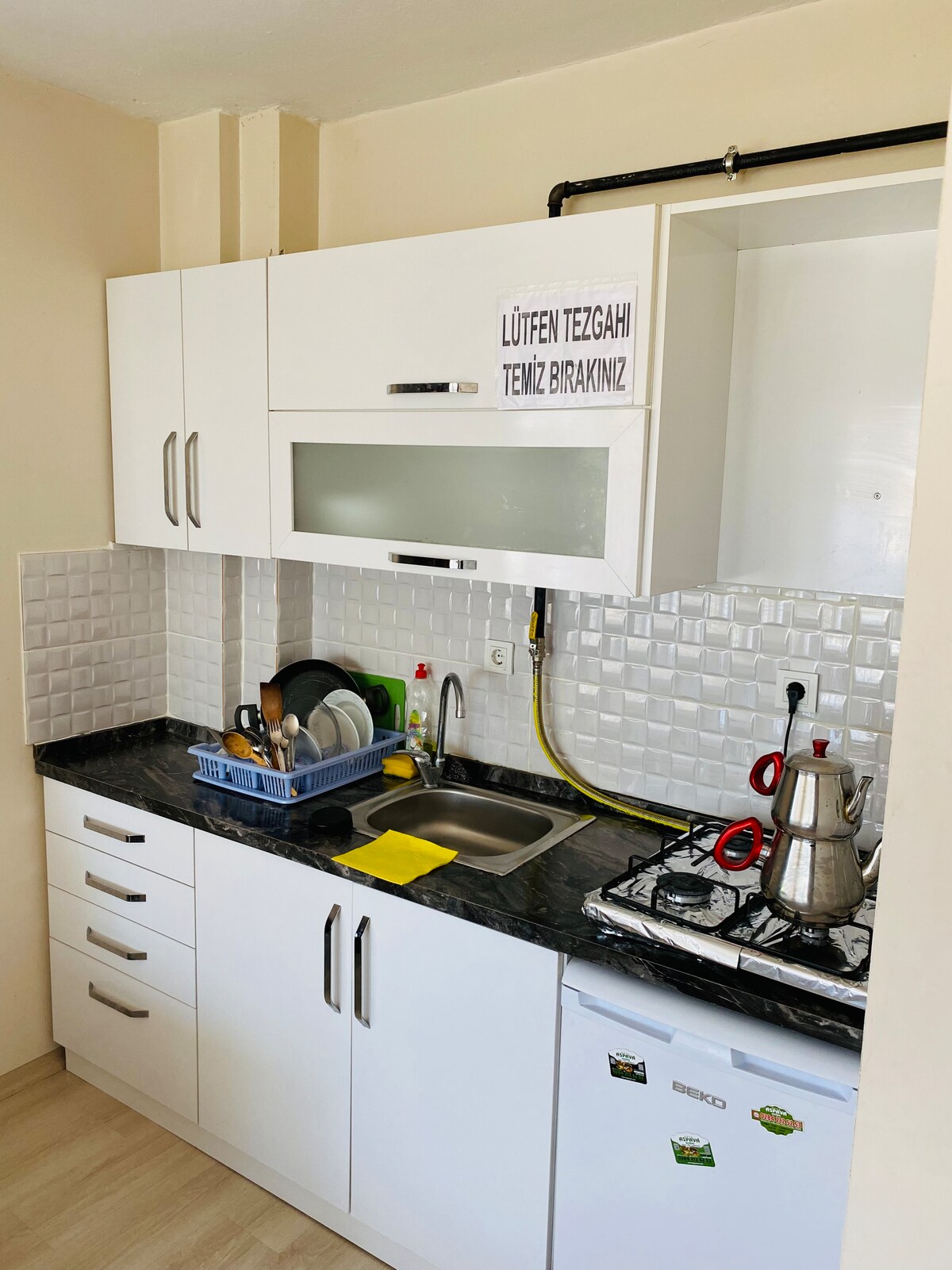 A compact kitchen features white cabinetry and a dark countertop. Various kitchen utensils are neatly organized on the countertop, with a sink and a stovetop visible. A small refrigerator is positioned beneath the counter, contributing to the functional layout.