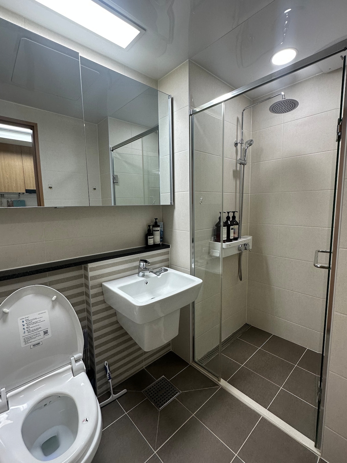 A modern bathroom features a glass-enclosed shower with a rain showerhead and a wall-mounted sink. A sleek toilet is positioned alongside a large mirror, reflecting the neutral tile design. Amenities such as toiletries are neatly arranged on the shelf.