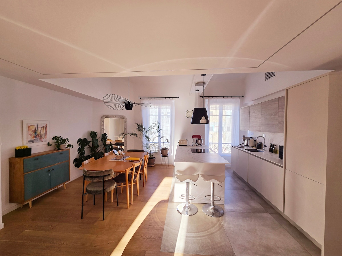 A modern kitchen area featuring a sleek island with bar stools and a dining table surrounded by chairs. Natural light streams through large windows, illuminating the space. A variety of plants add a touch of greenery, enhancing the open design.