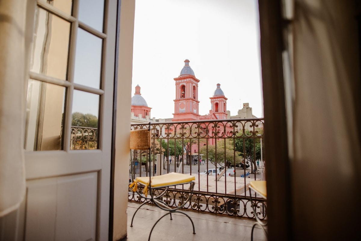 A balcony offers a view of two pink towers rising above the surrounding landscape. The railing is intricately designed, and a pair of yellow lounge chairs is positioned on the balcony, inviting relaxation while enjoying the scenery.
