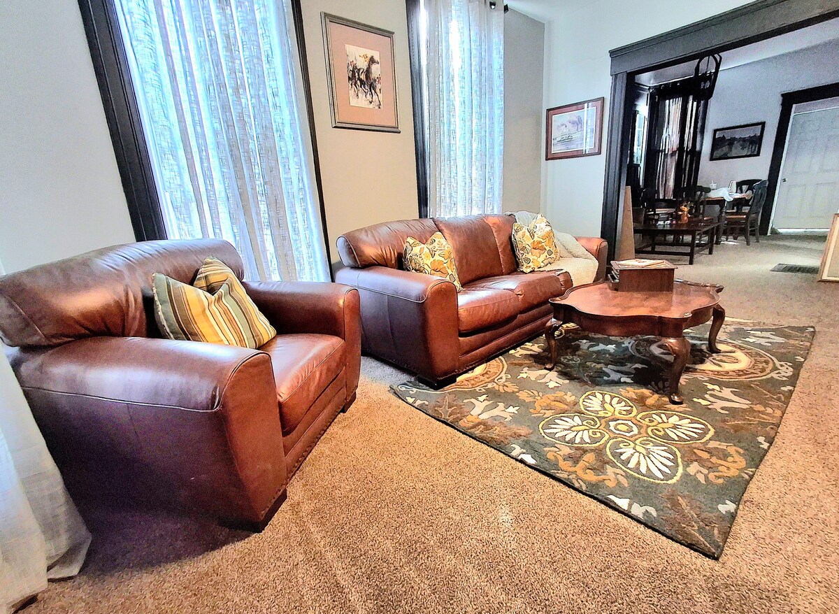 A spacious living room features a leather couch and two matching chairs, accented with decorative pillows. A central coffee table rests on a patterned rug, while tall windows, adorned with sheer curtains, allow natural light to fill the room.