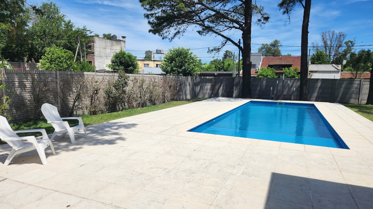 A spacious outdoor area features a rectangular swimming pool surrounded by a tiled patio. Two white lounge chairs are positioned beside the pool, set against a backdrop of greenery and tall trees. The serene environment offers a sense of openness and tranquility.