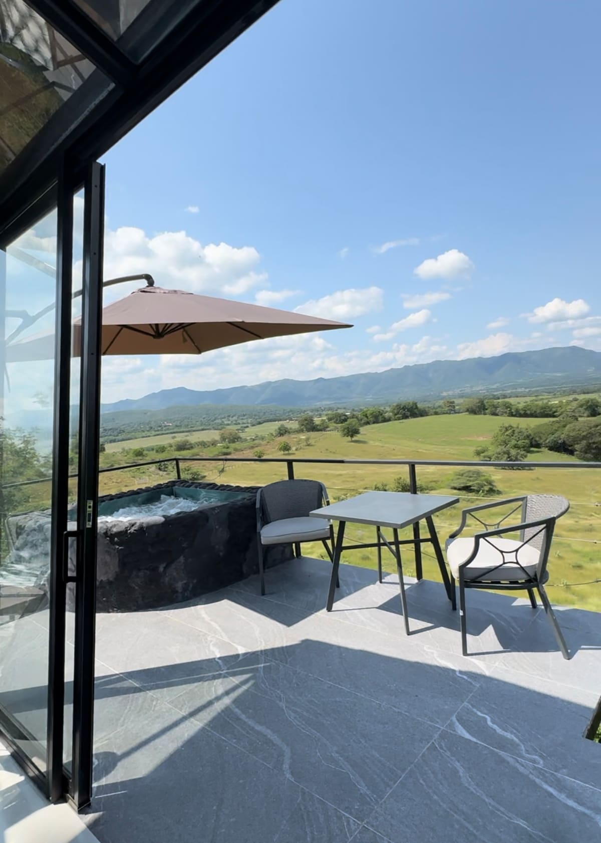A patio area features a square table and four chairs beneath an umbrella. A hot tub is positioned next to the railing, offering views of expansive green fields and distant mountains under a bright blue sky with scattered clouds.
