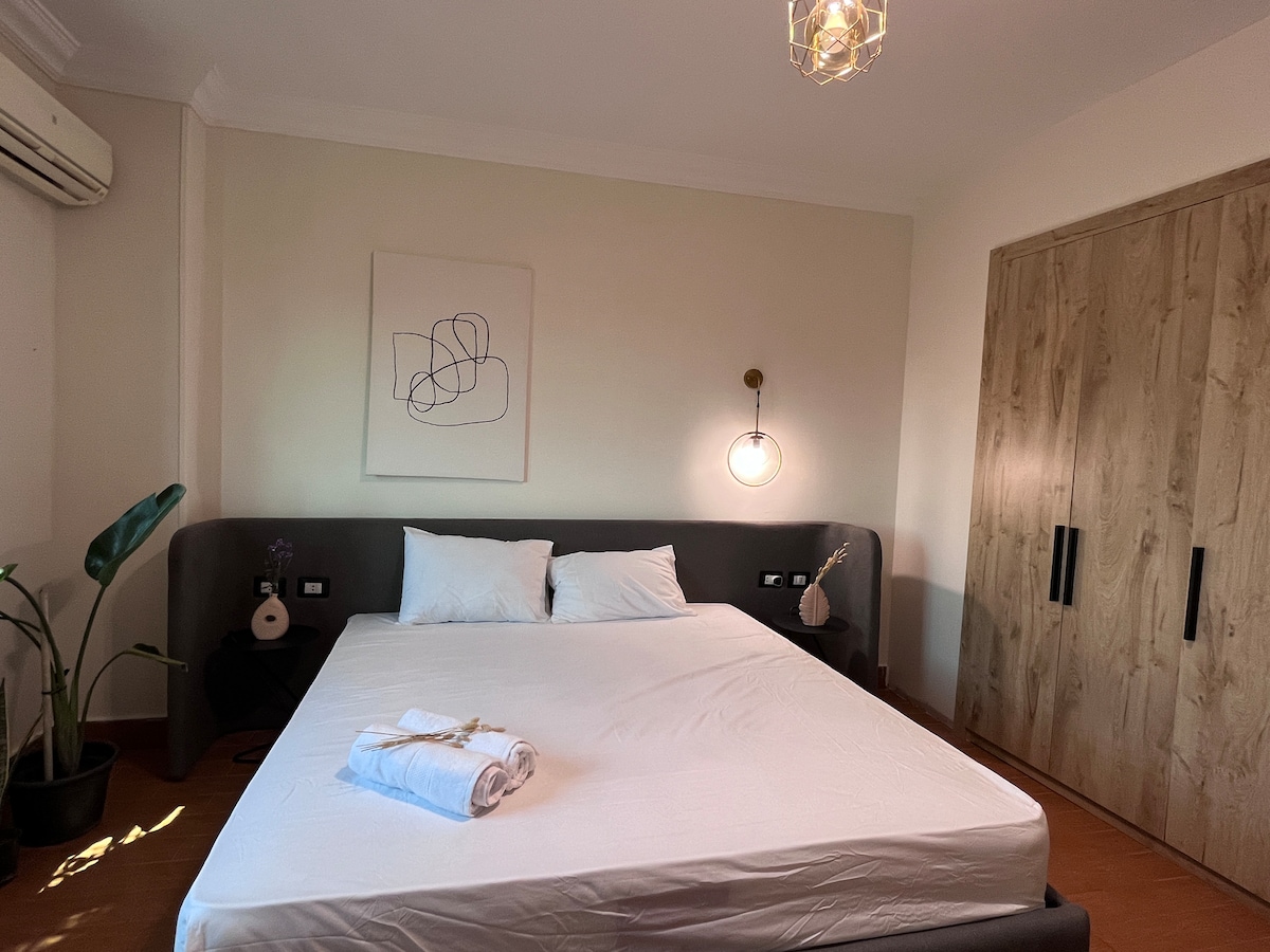 A bright bedroom features a spacious bed with crisp linens and two soft pillows. A minimalist wall art piece is displayed above the bed, accompanied by a modern pendant light. A leafy plant adds a touch of nature, and a wooden wardrobe provides storage.