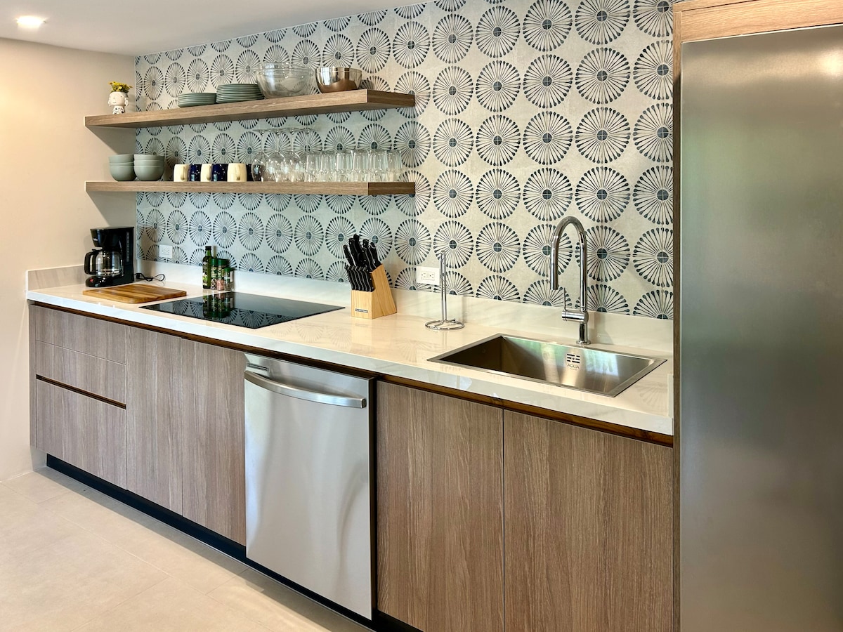 A modern kitchen is showcased, featuring wooden cabinetry and a sleek countertop. Open shelves display dishware against a patterned backsplash. Stainless steel appliances include a refrigerator and a sink, with utensils neatly arranged in a wooden holder, contributing to a functional cooking space.