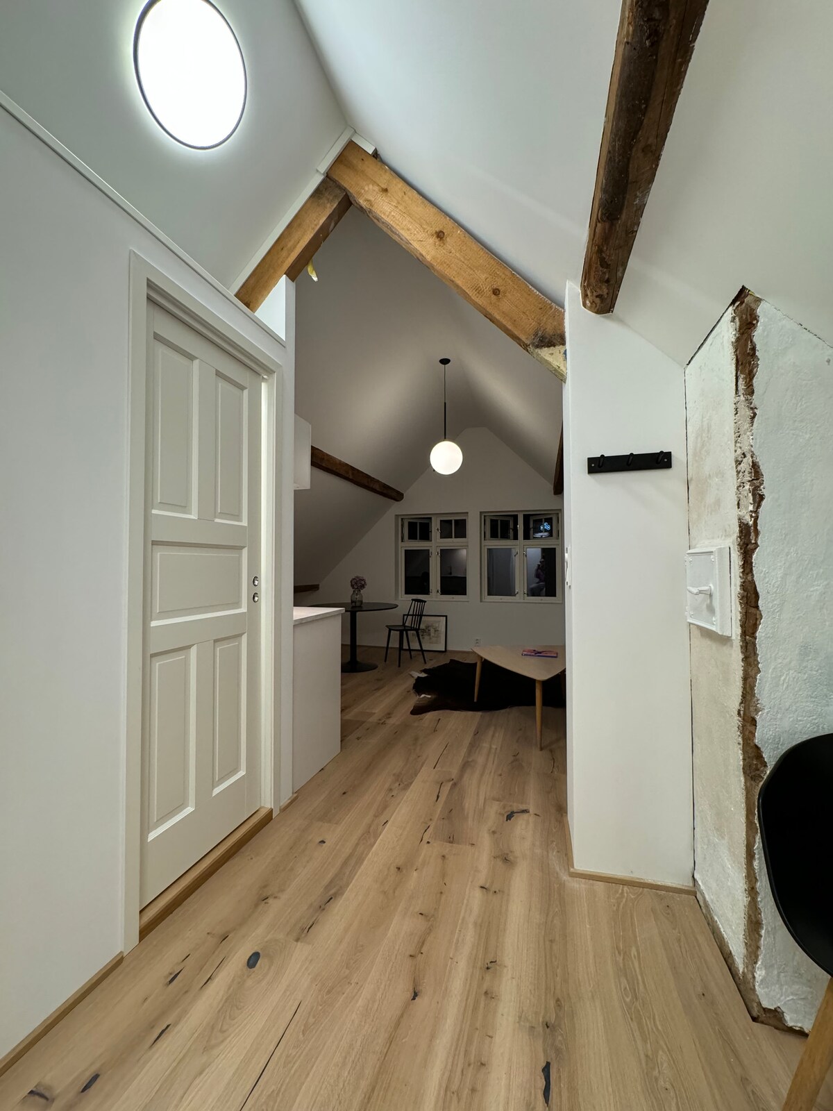 A hallway with high ceilings is displayed, featuring wooden beams that add architectural interest. A round ceiling light provides illumination. A smooth wooden floor extends towards a cozy seating area visible in the background, complemented by a simple table and modern decor.